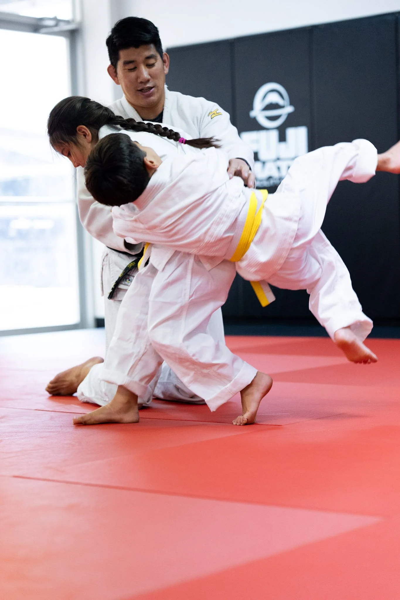 Two children practicing judo on a red mat with an instructor assisting, wearing white gi uniforms with yellow belts.