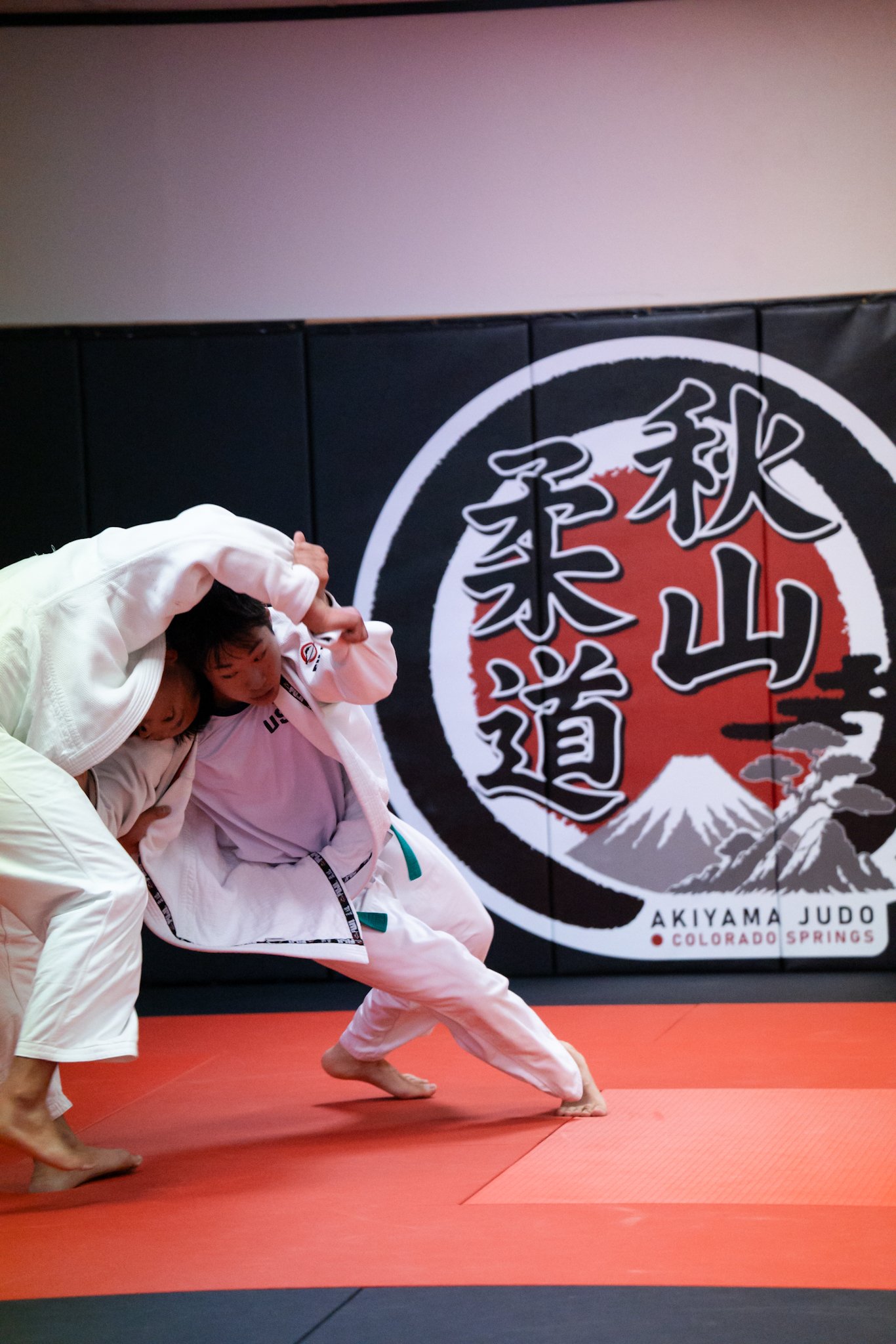 Two children practicing judo on an orange mat at Akiyama Judo in Colorado Springs with a large black and red logo of Mount Fuji and Japanese characters on the wall in the background.