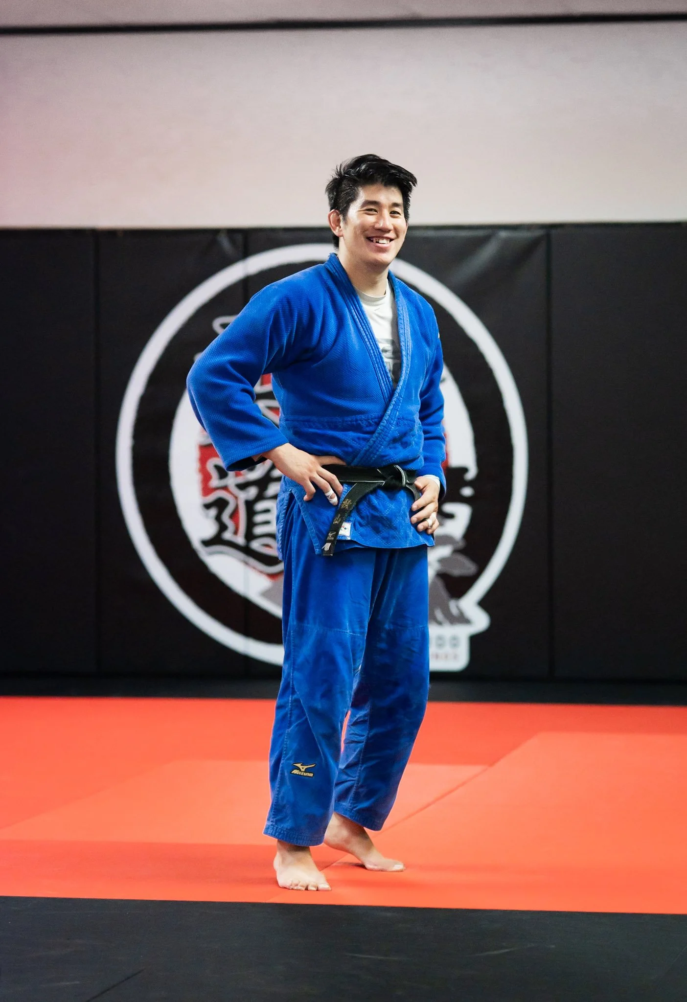 A young man with black hair wearing a blue Brazilian judo gi standing on a red and black judo mat, smiling and resting his right hand on his hip. There is a logo on his pant leg and a black background with a white circular symbol behind him.