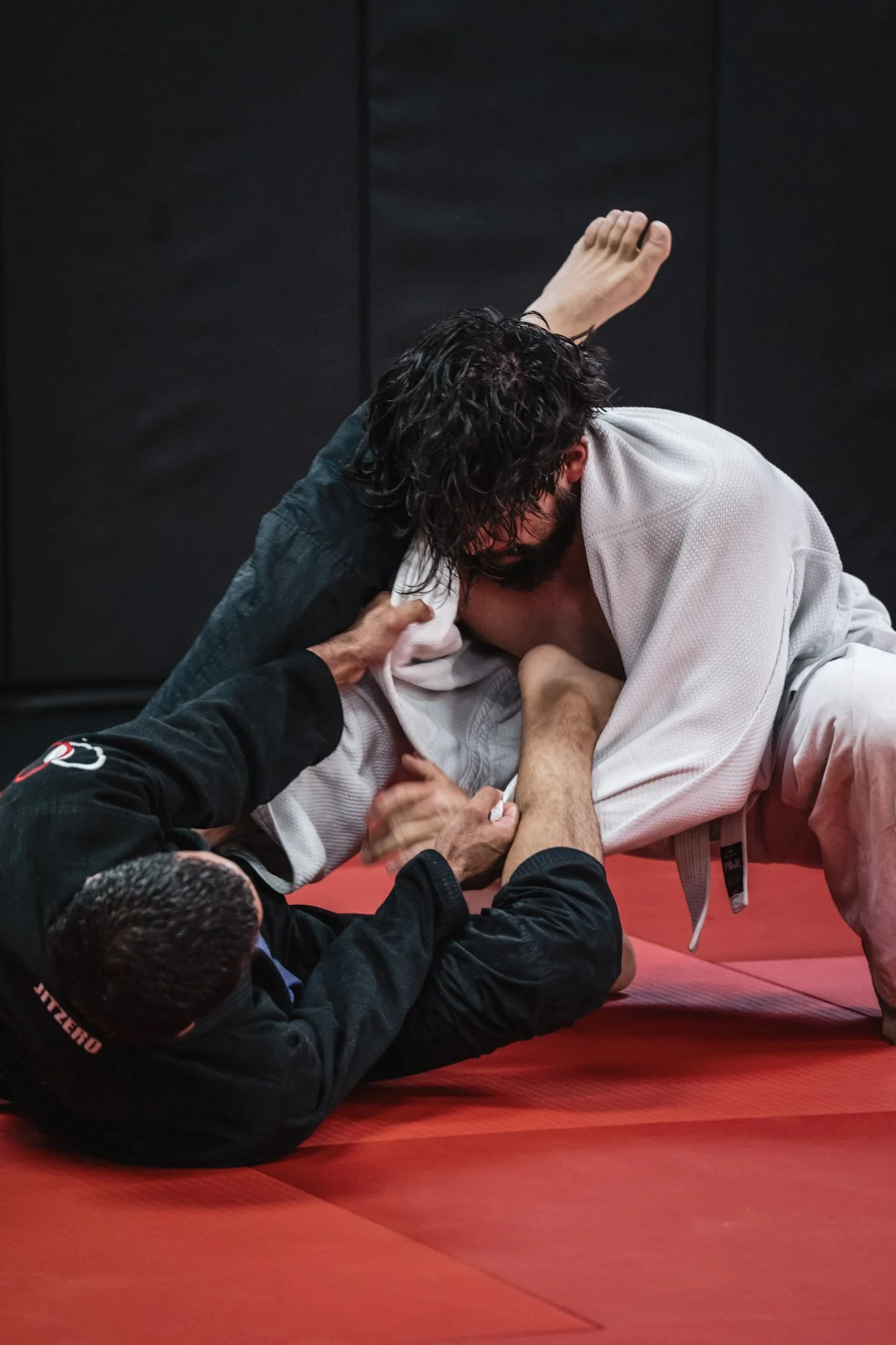 Two individuals practicing Brazilian Jiu-Jitsu on red mats, one wearing a white gi and the other a black gi, engaging in a close grappling position indoors.