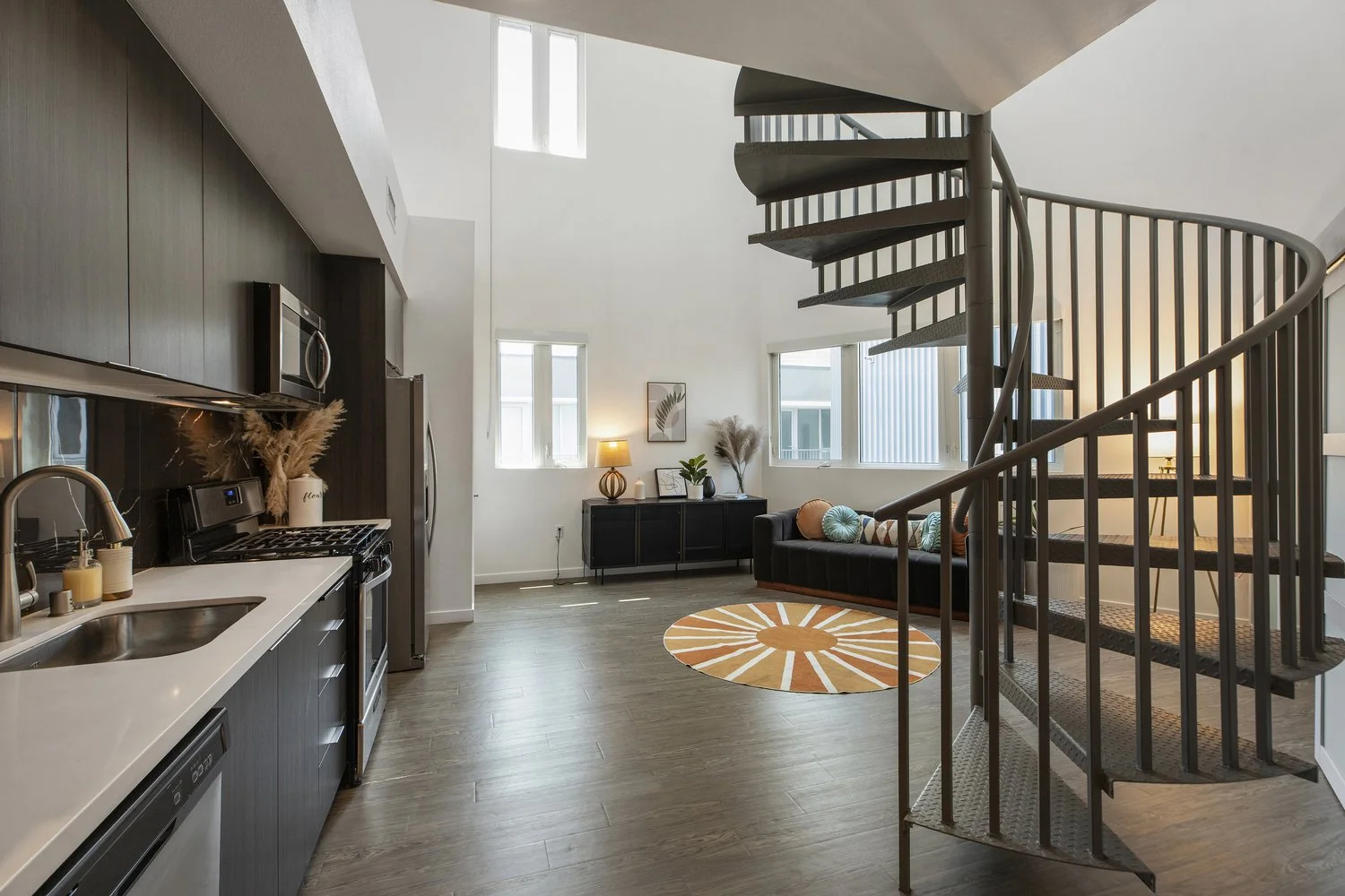Open-concept living room and kitchen with a spiral staircase, black sofa with decorative pillows, round orange and white rug, and large windows allowing natural light.