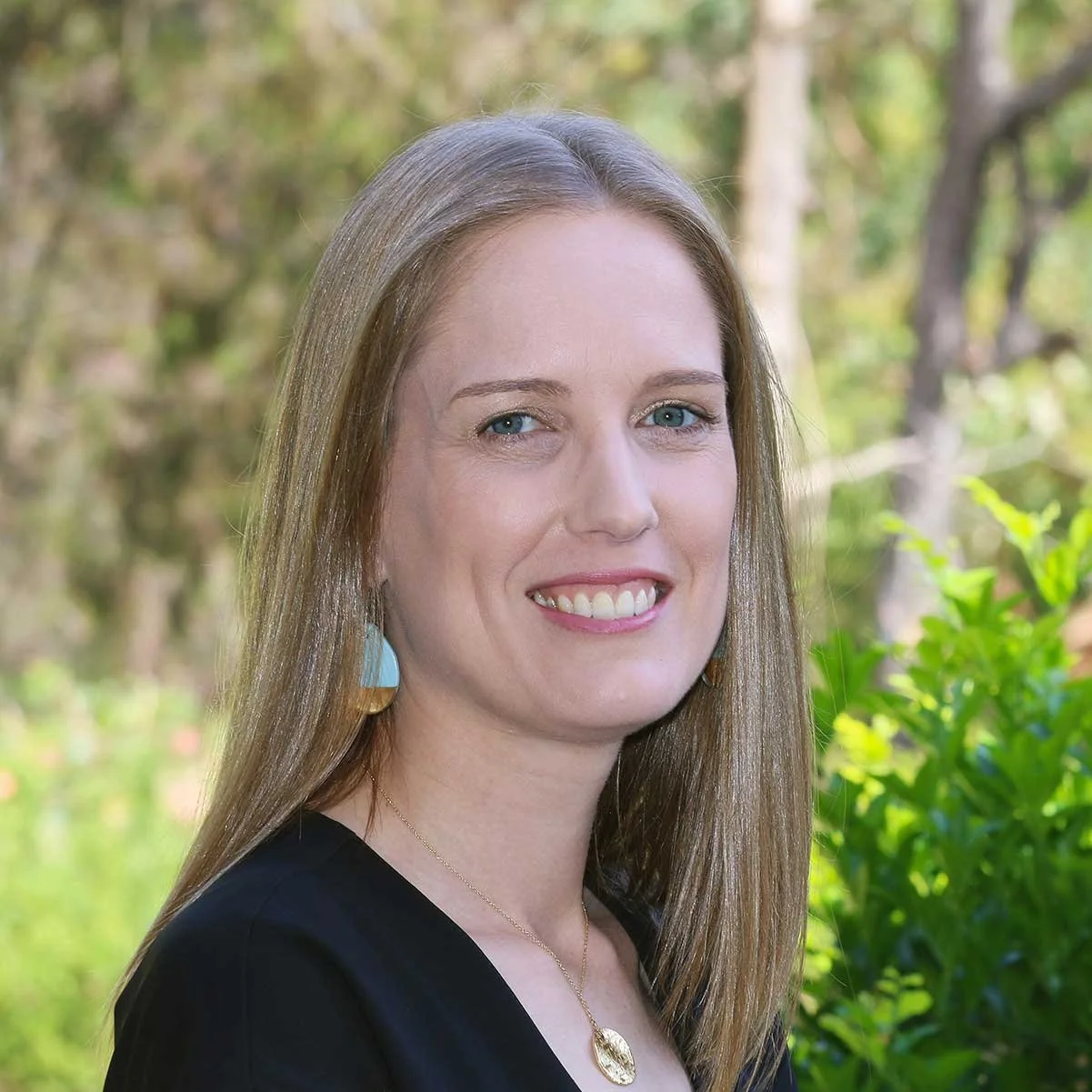A woman with long light brown hair and blue eyes smiling outdoors with greenery and trees in the background.