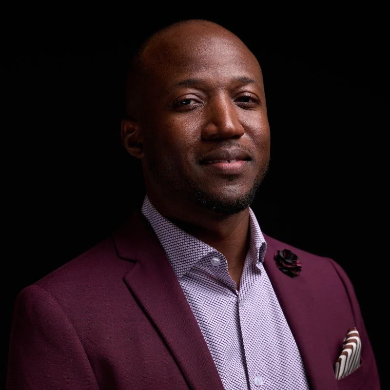 Portrait of a confident African American man in a maroon suit and light patterned shirt against black background.