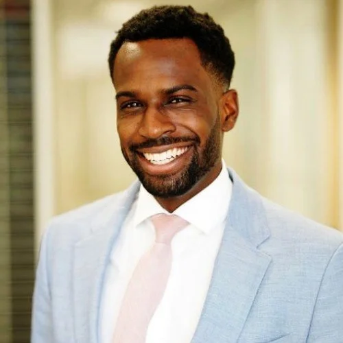 Headshot of a smiling African American man in a light gray suit and pink tie, standing outdoors.