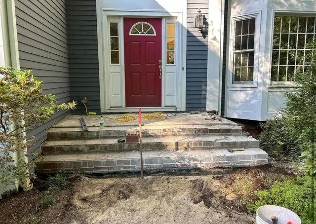 Front steps of a house under renovation with brick work in progress, a red front door, and surrounding house siding and windows.