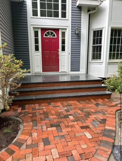 Red front door with white frame and small window, three black steps leading to a gray porch, brick pathway, and surrounding greenery.