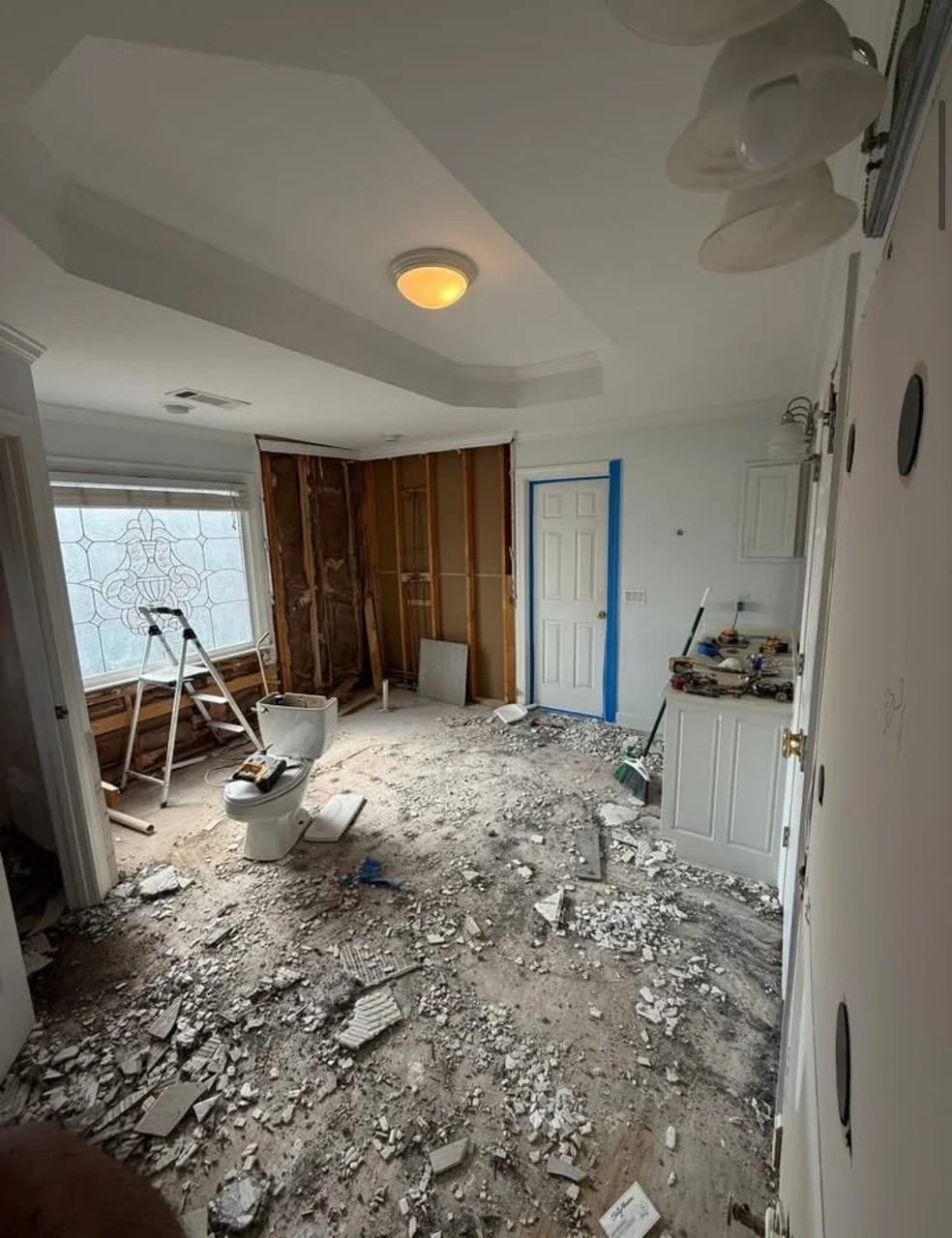 A room under renovation with stripped walls, exposed wall studs, and debris on the unfinished floor. There is a toilet and a ladder near a window with decorative glass. Construction tools and supplies are scattered around.