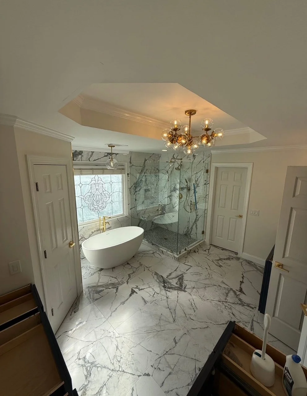 Elegant bathroom with marble floors, freestanding bathtub, glass-enclosed shower, chandelier, and a frosted decorative window.