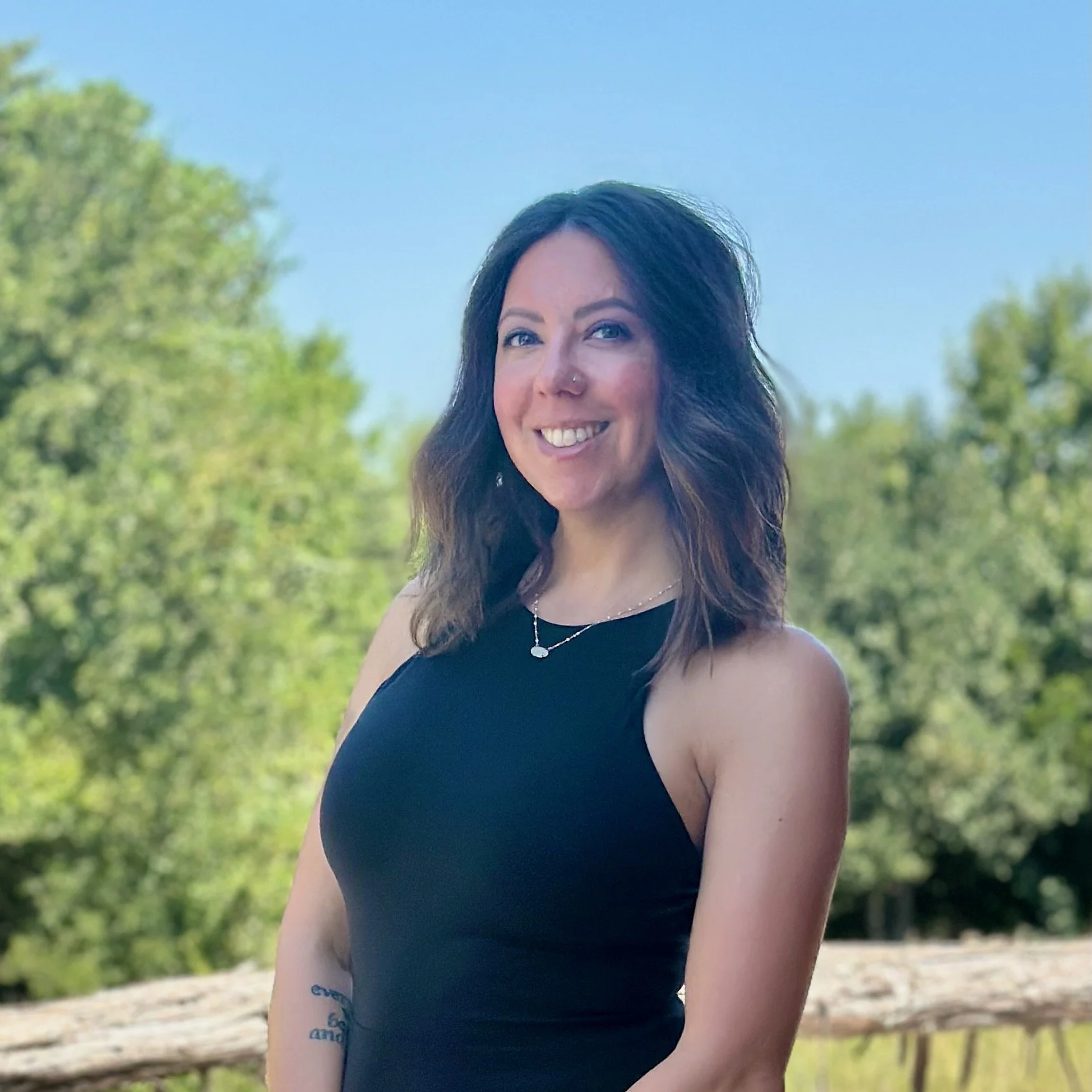 A woman with dark hair, wearing a black sleeveless top, smiling outdoors with greenery and blue sky in the background.
