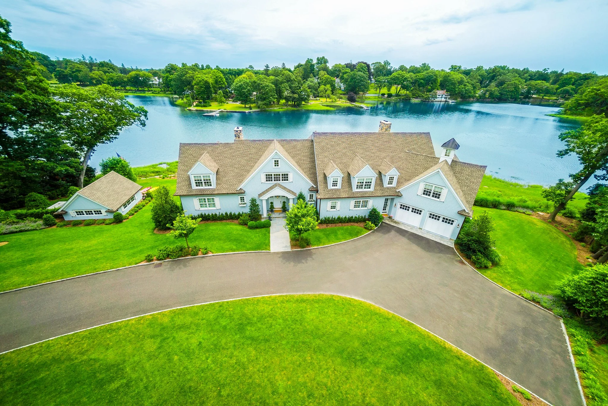 A large house with white siding and multiple gabled roofs is situated on a lush green lawn next to a curved driveway. The property overlooks a body of water with several small boats and an expansive view of a wooded area and additional houses across the water.