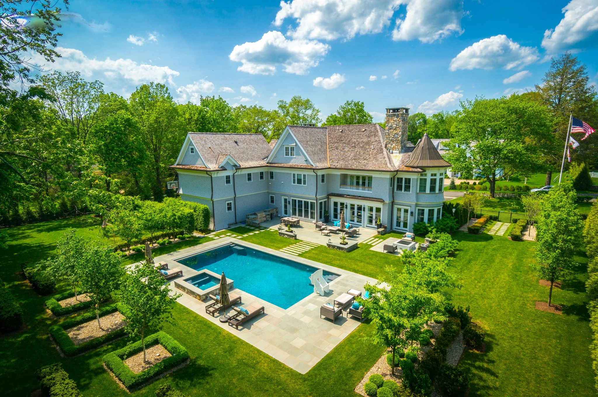 A large, elegant house with a swimming pool in the backyard, surrounded by lush green trees and a well-manicured lawn under a bright blue sky with some clouds.