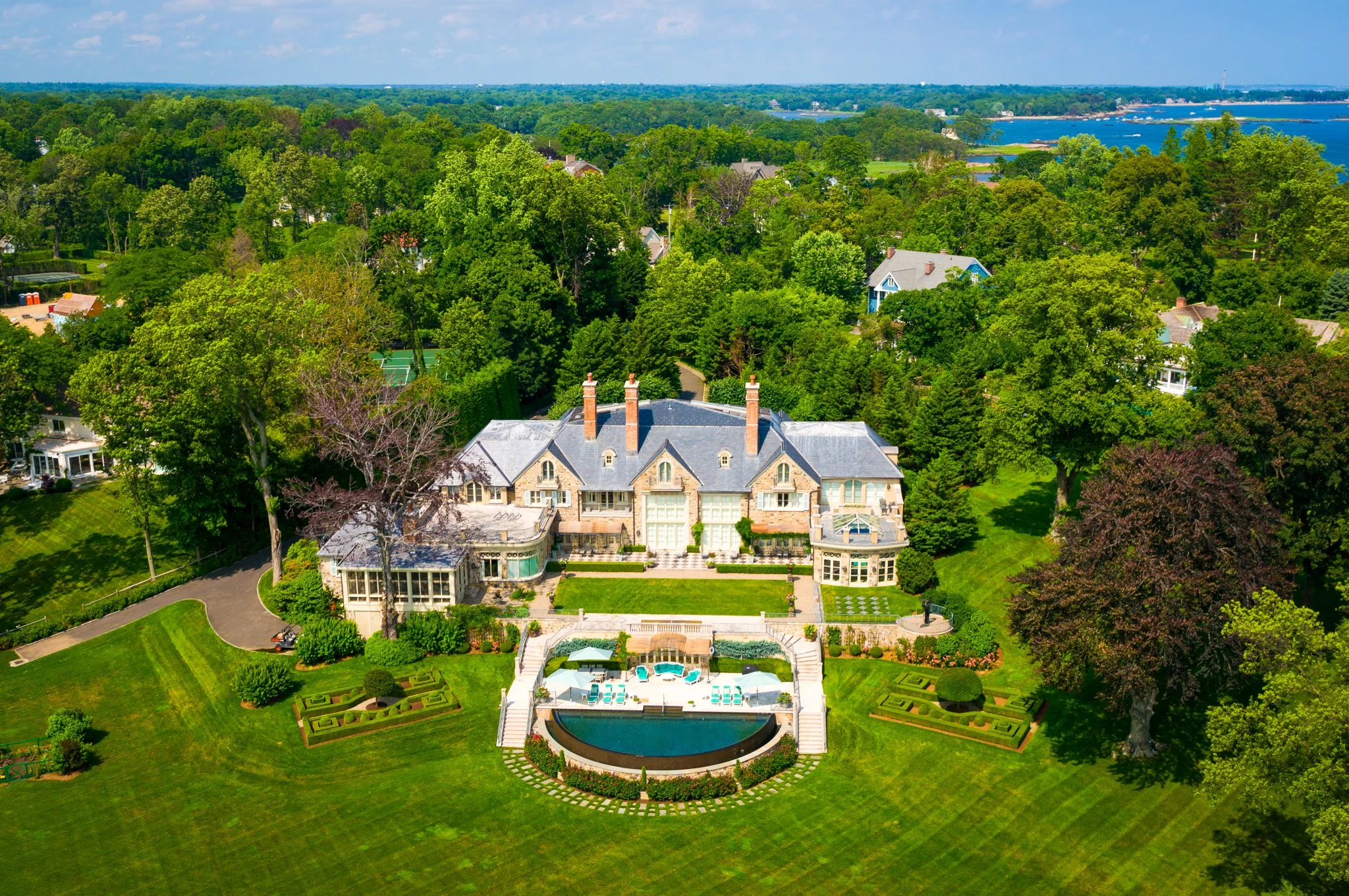 An aerial view of a large house with a gray roof, surrounded by lush green trees, a well-maintained lawn, and a backyard with a swimming pool and patio area, with a waterfront and city skyline in the distance.