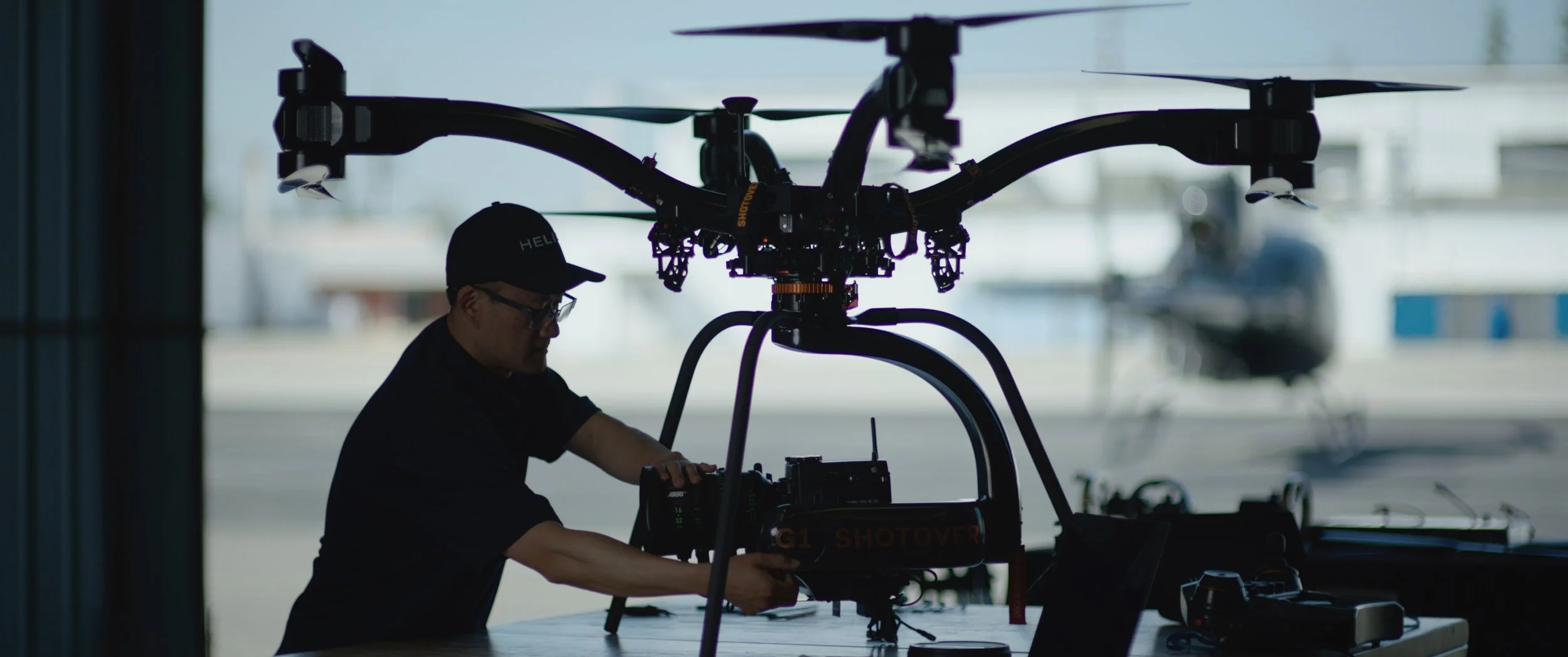 Person working on a large drone inside a hangar with blurred aircraft outside in the background.