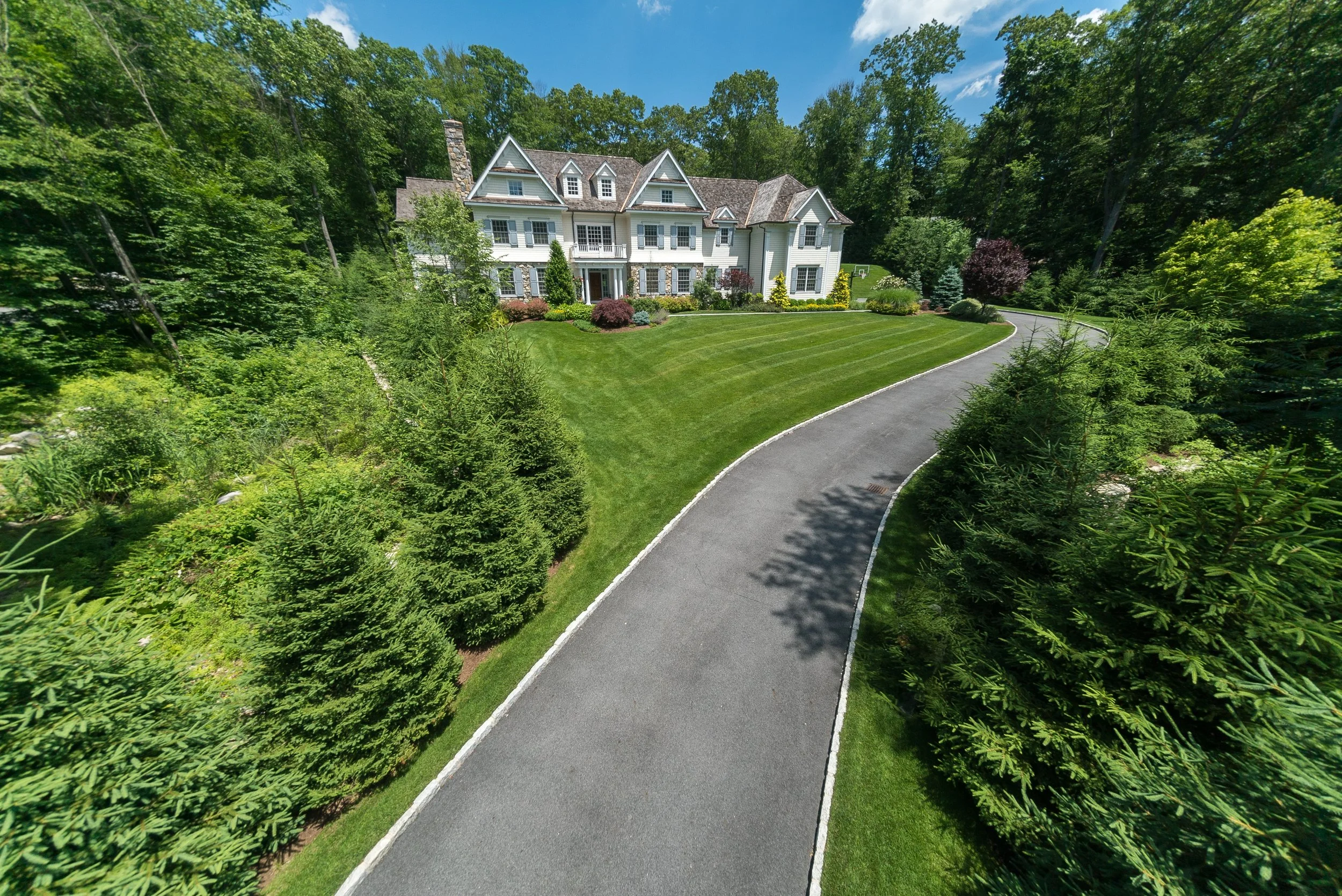 A large white house with multiple steep gabled roofs and a stone chimney, surrounded by a lush green landscape with manicured lawn and a curved driveway.
