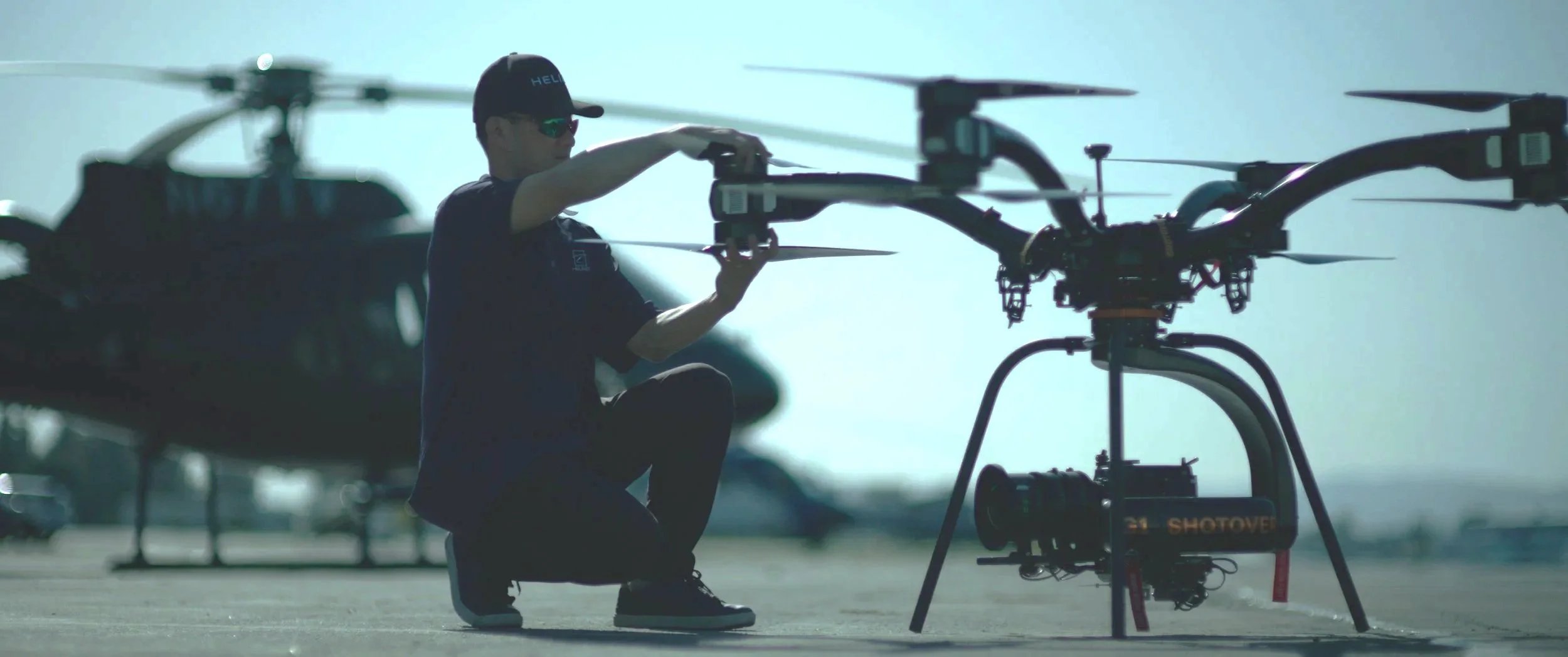A technician working on a large drone with helicopters in the background on an airfield.