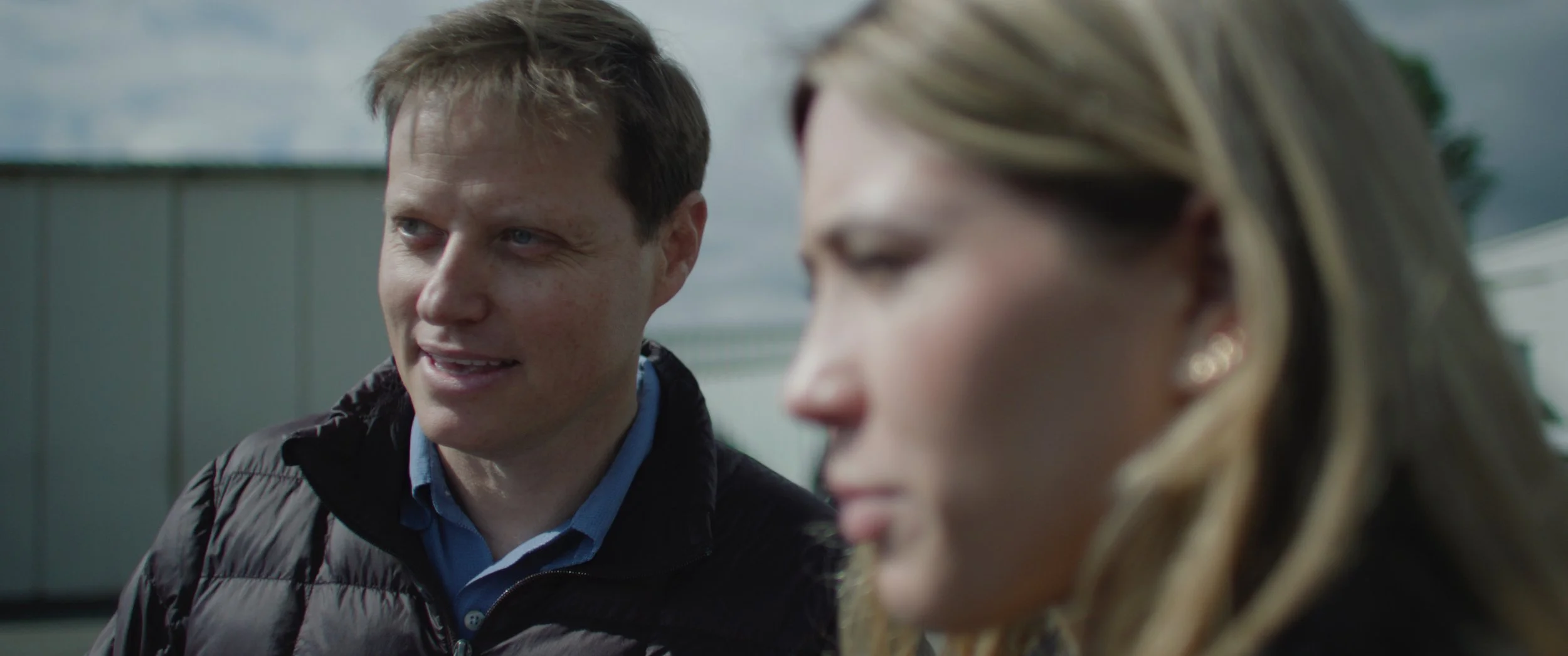 A man with short brown hair and a woman with long blonde hair and earrings are outdoors, engaged in conversation. The man is smiling and dressed in a jacket and collared shirt.