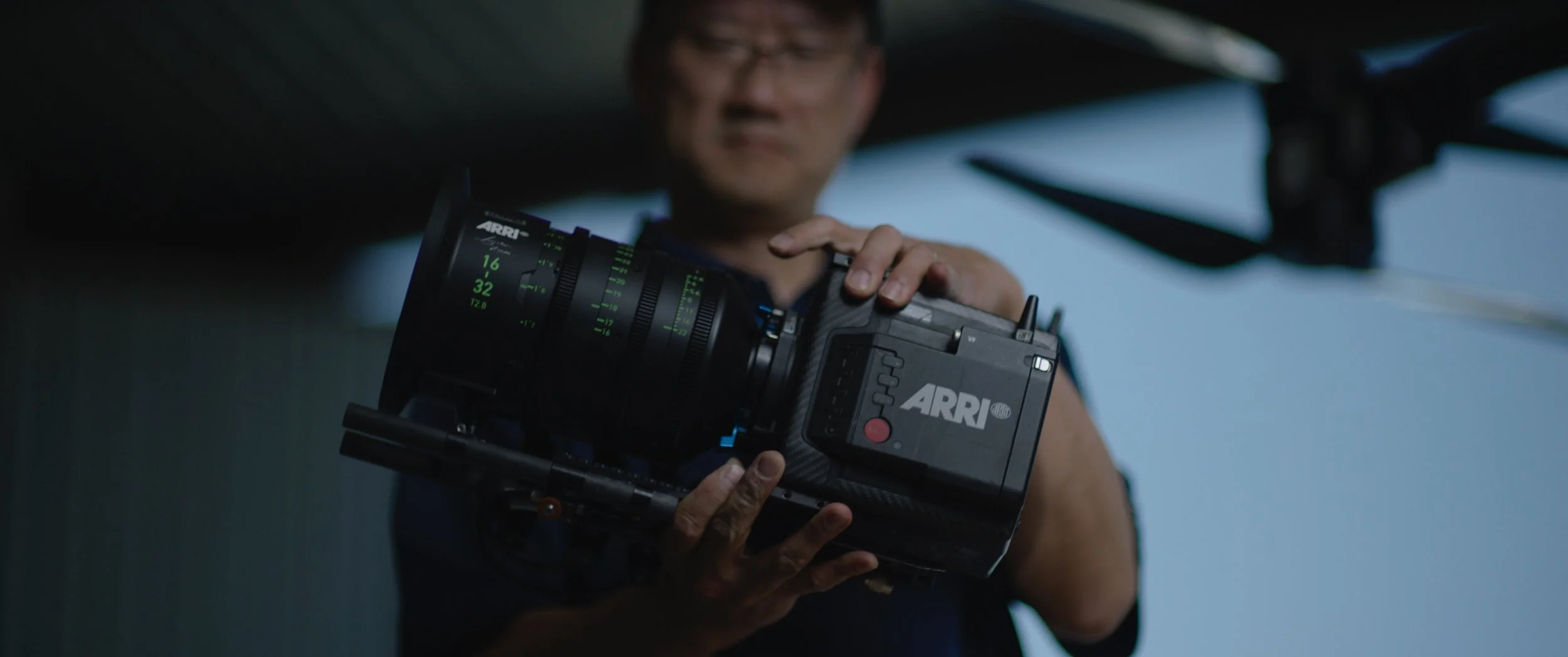 A person holding an ARRI professional camera, adjusting settings on a film or video shoot under a structure with a background of a clear blue sky.