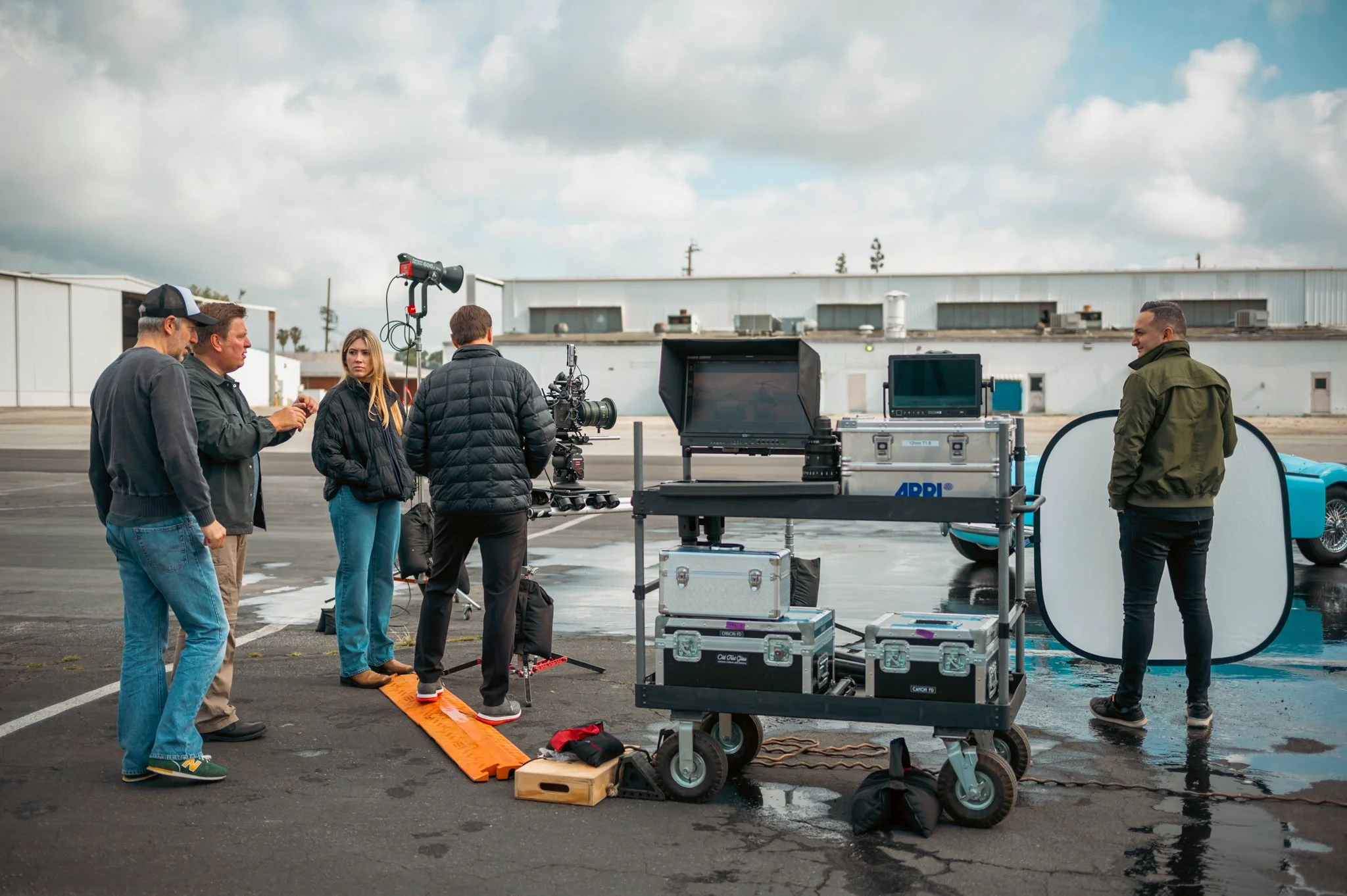 Film crew setting up equipment outside on a cloudy day, including cameras, monitors, and lighting, near a white industrial building.