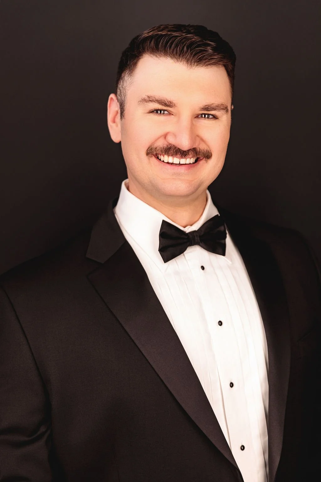 Chris Gallerani - Tenor, smiling in a tuxedo with a black bow tie and a white dress shirt against a dark background. Headshot by Benjamin Rose Photography.