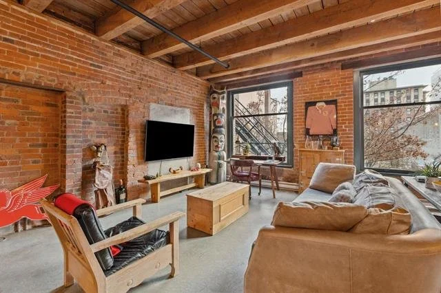 Living room with exposed brick walls, large windows, a beige sofa, a wooden armchair with a black cushion, a flat-screen TV on the wall, and a wooden coffee table.