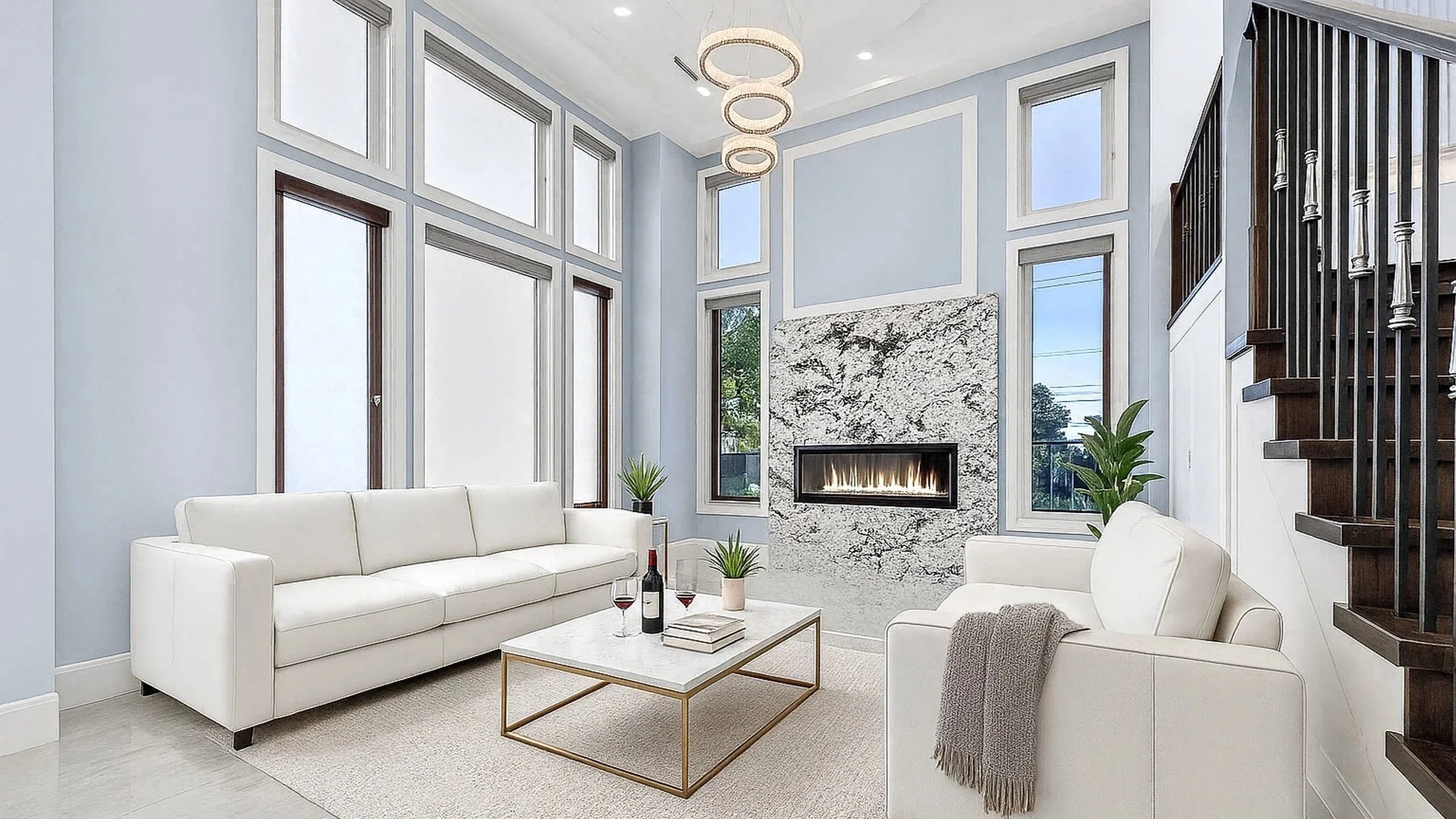 Living room with light blue walls, tall narrow windows, a modern fireplace with a stone surround, white sofas, a coffee table with a bottle of wine and wine glasses, and plants near the windows.