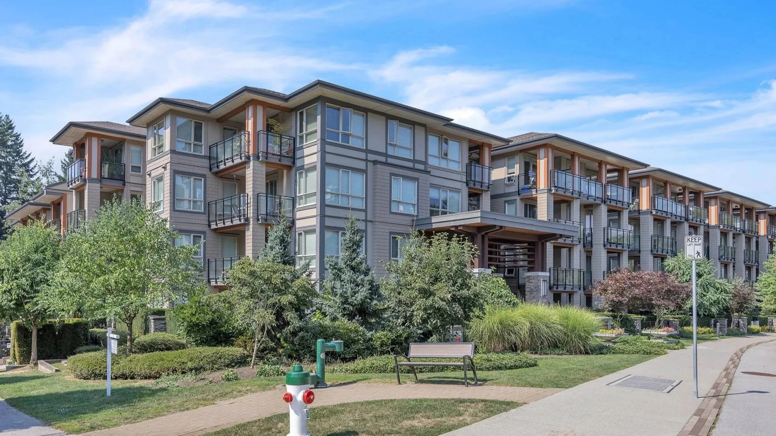 A modern multi-story apartment building with balconies, surrounded by lush trees and greenery, with a clear blue sky in the background.