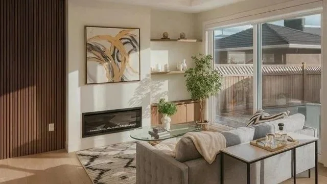 Modern living room with a gray sofa, glass coffee table, abstract art on the wall, built-in fireplace, potted plant, and large sliding glass door leading to a backyard.