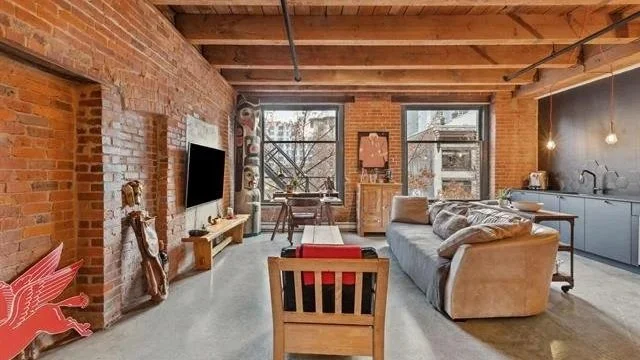 Open-concept living room in a loft with exposed brick walls, large windows, a beige sectional sofa, a wooden armchair, a TV mounted on the brick wall, and a modern kitchen with blue cabinets in the background.