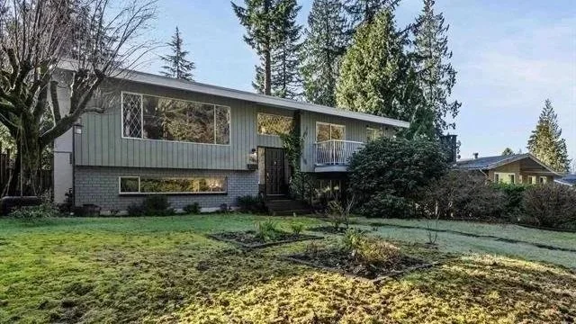A mid-century modern house with gray siding, large windows, and a small front yard with-green grass, trees, and shrubs, situated in a suburban neighborhood.