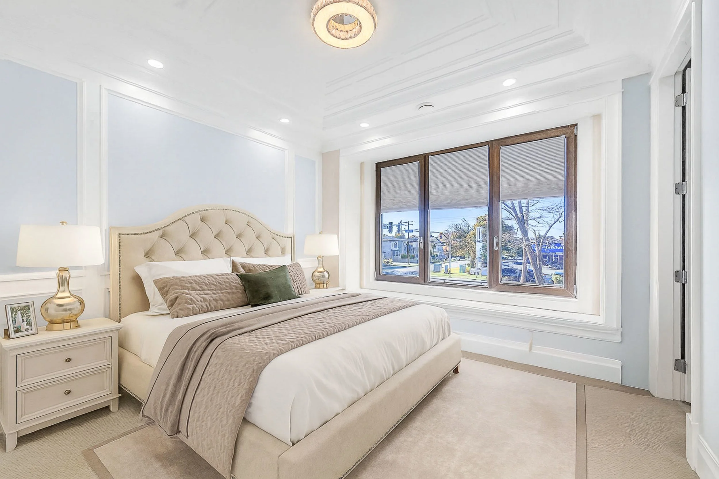 A bedroom with a large bed featuring a tufted headboard, neutral bedding, and decorative pillows. There are two matching nightstands with lamps and framed photos. The room has large windows with blinds, white walls with molding, a ceiling light fixture, and is carpeted.