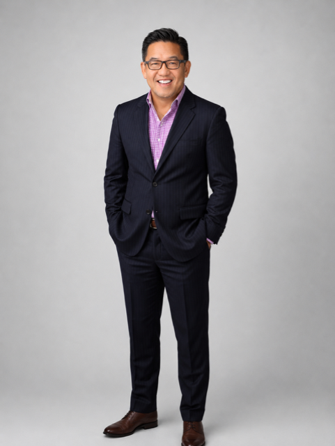 A smiling man wearing glasses, a dark suit, and a purple checkered shirt standing against a plain light gray background.