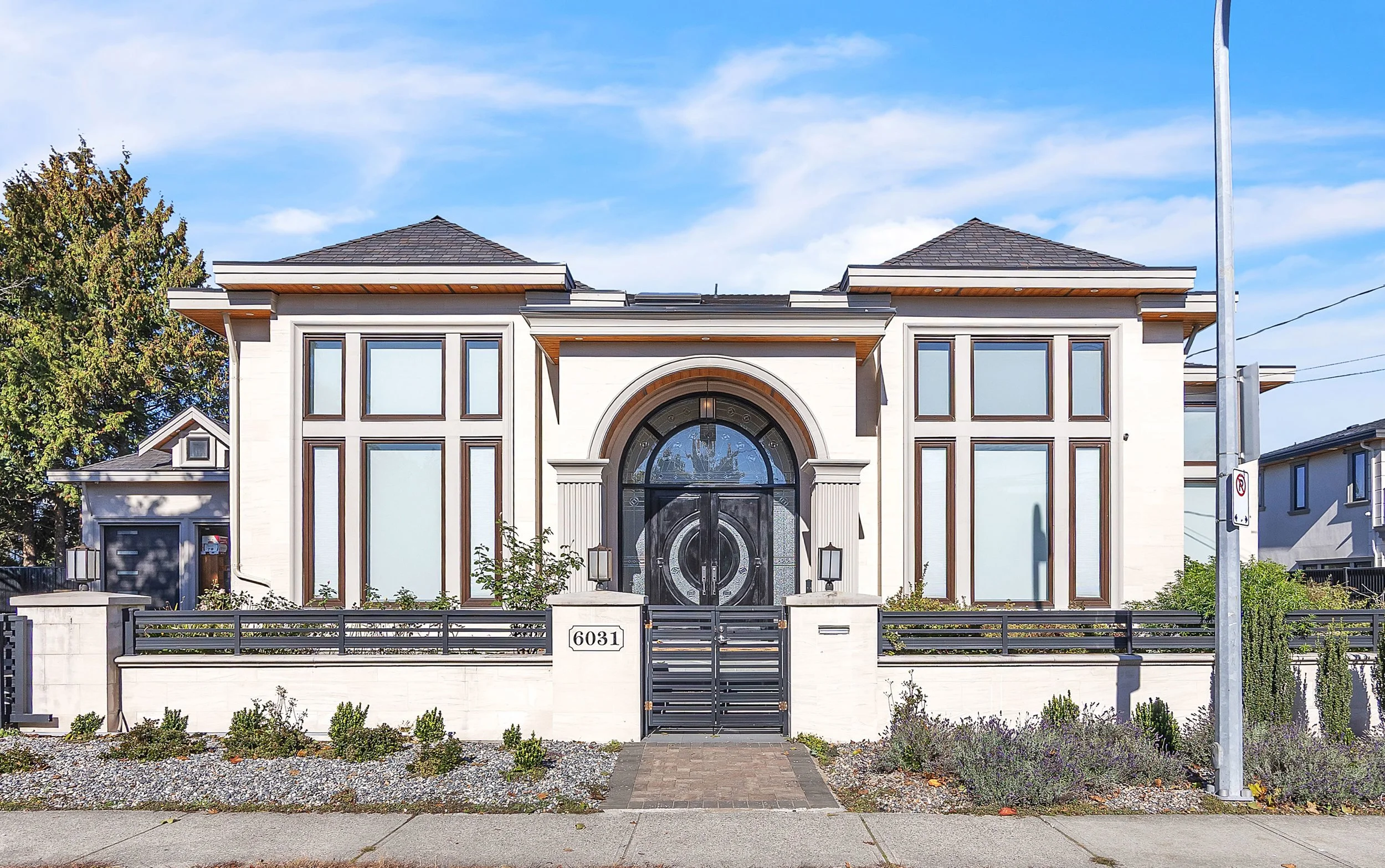 Modern two-story house with white exterior, large arched front door, and multiple windows, enclosed by a metal fence, with a sidewalk and well-maintained landscaping in the front yard. The house number '6031' is on the gate post.