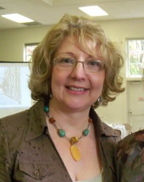 A woman with shoulder-length blonde hair, wearing glasses, a brown blouse, and a colorful necklace, smiling indoors.