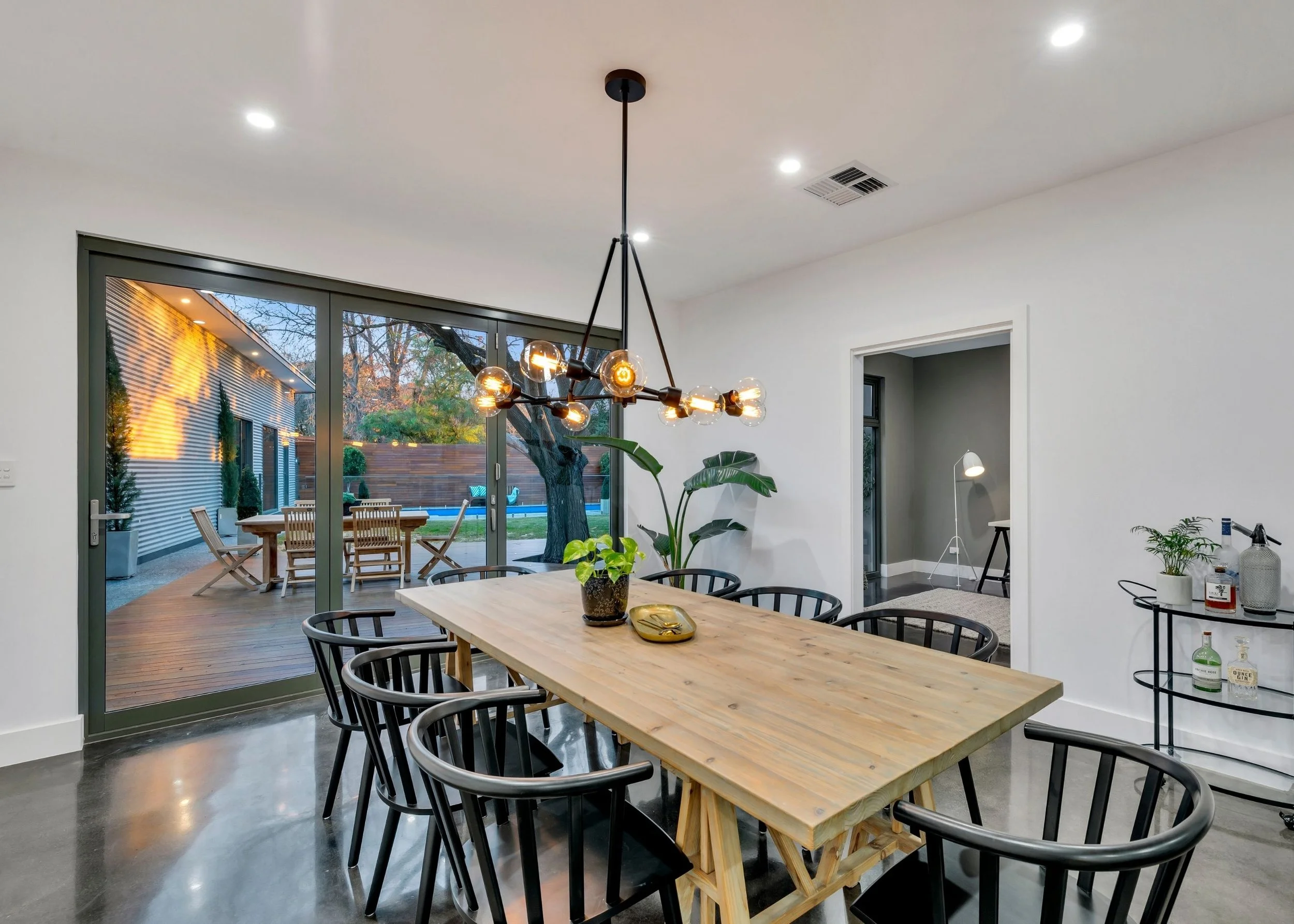 Modern dining room with a large wooden table surrounded by black chairs, a contemporary chandelier, and large sliding glass doors opening to an outdoor patio with a wooden dining set and trees.