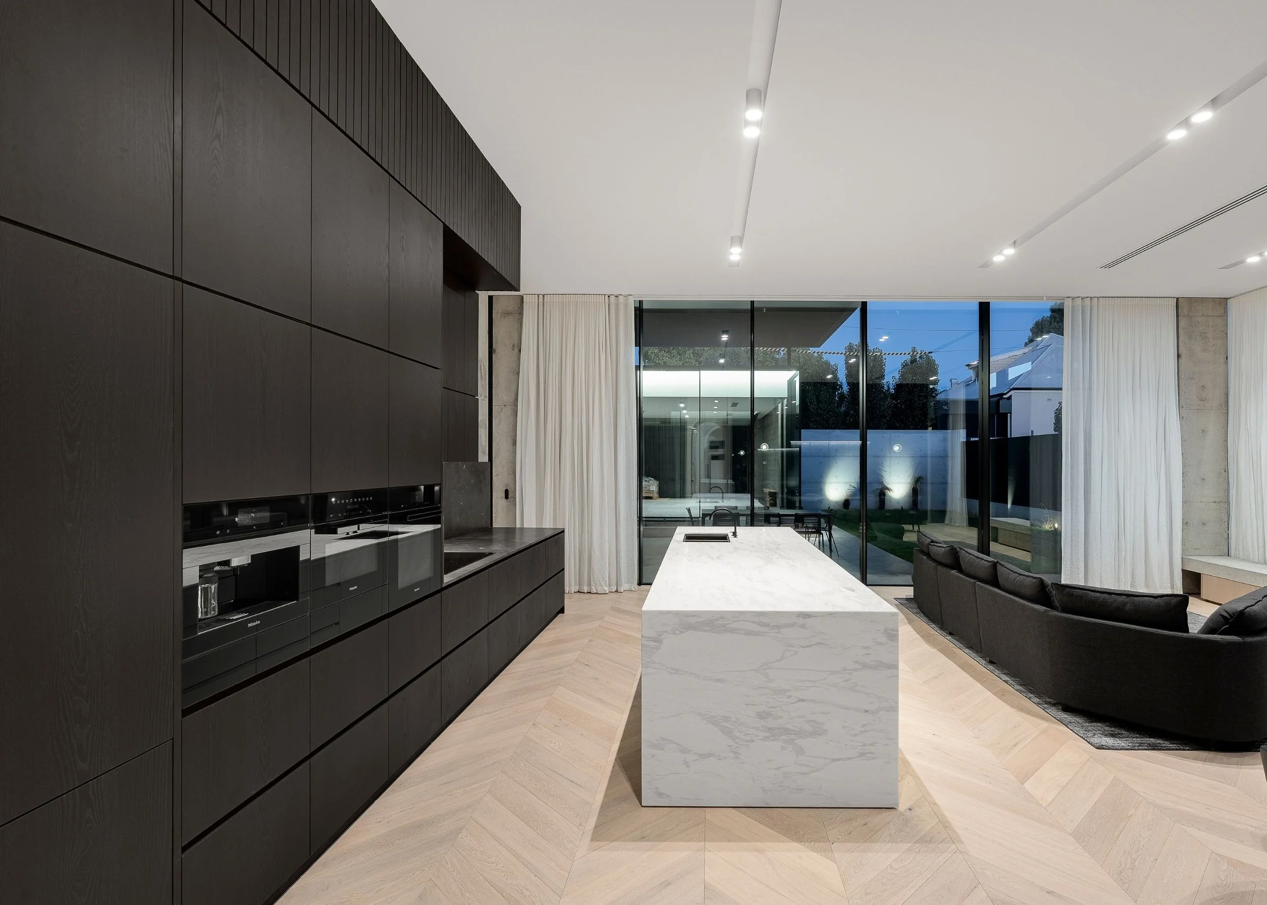 Modern kitchen and living area with dark cabinetry, white marble island, large windows with white curtains, and outdoor patio visible through the glass.