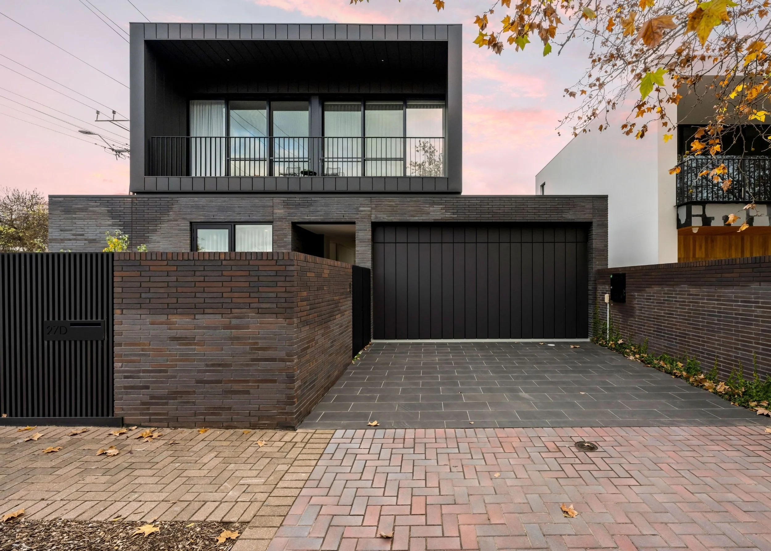 A modern multi-story house with dark exterior walls, large glass windows, and a balcony, surrounded by brick walls and a paved front yard, during autumn with fallen leaves and a colorful sky.