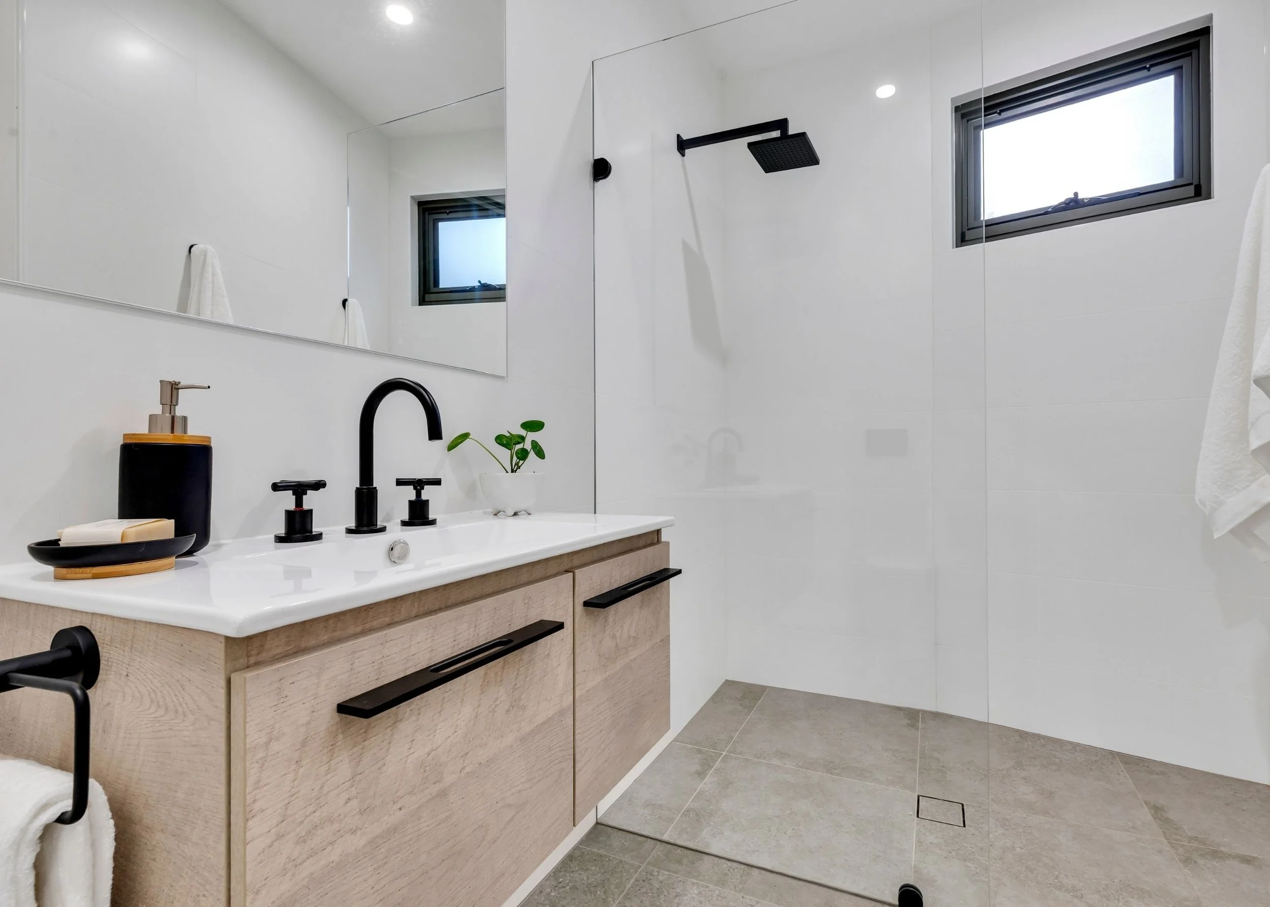 Modern bathroom with white walls, a large mirror, a black showerhead, a window, a wooden vanity with black handles, a plants, soap dispenser, and a towel.