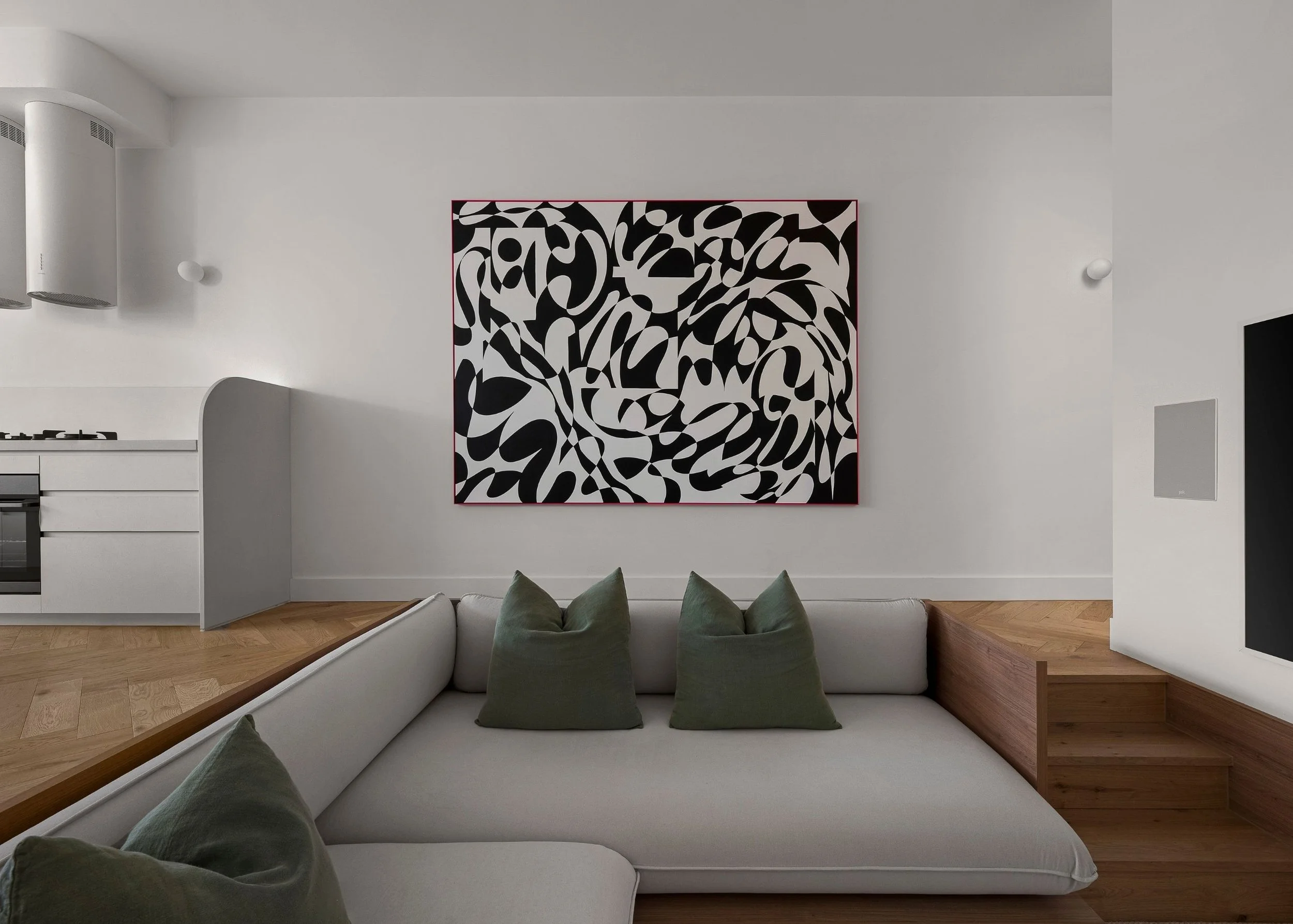 Modern living room with white sofa and green cushions, black and white abstract artwork on the wall, open kitchen in the background, hardwood flooring.