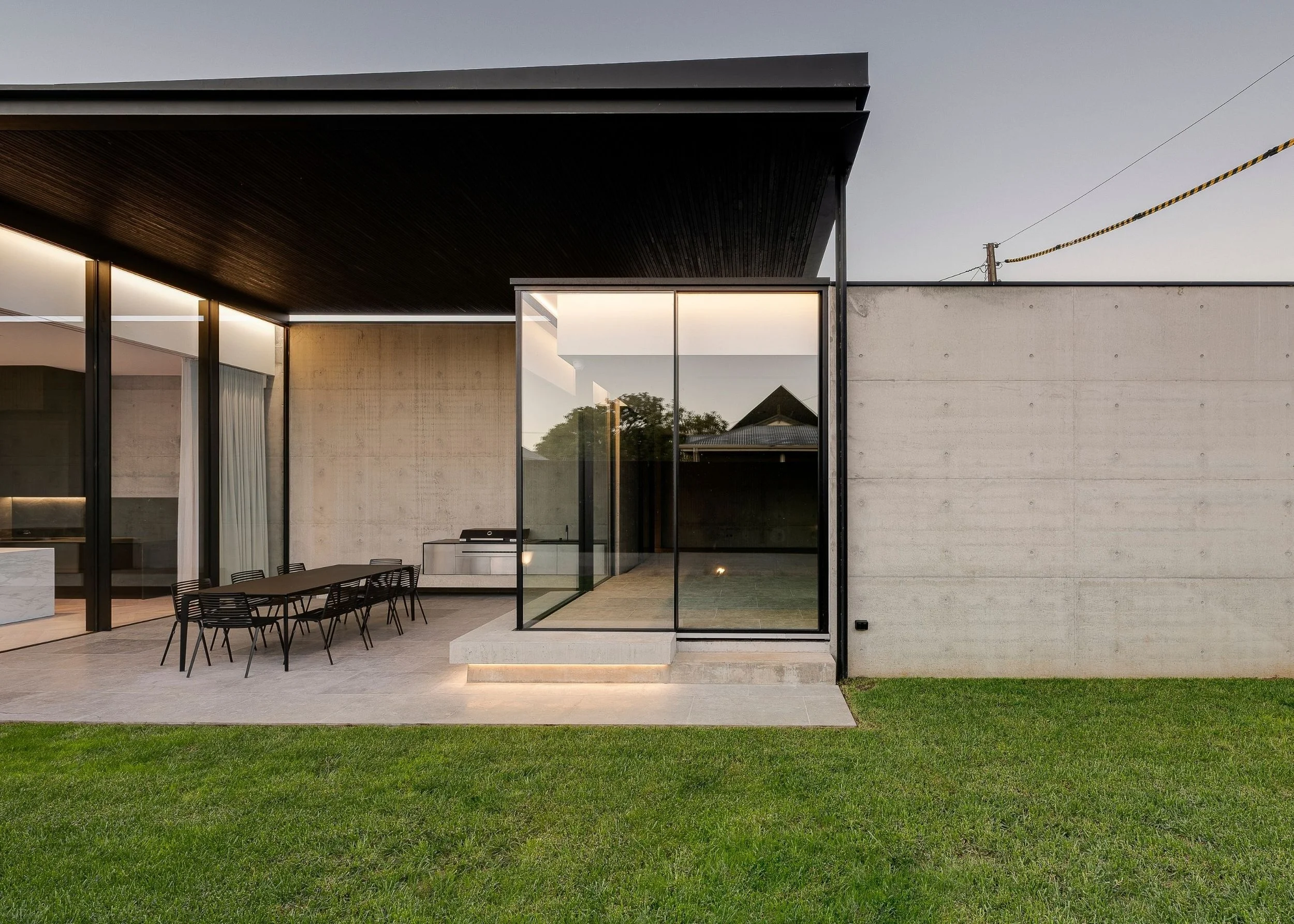 Modern house exterior with large glass windows, a small outdoor dining table with chairs, concrete walls, and a grass lawn.