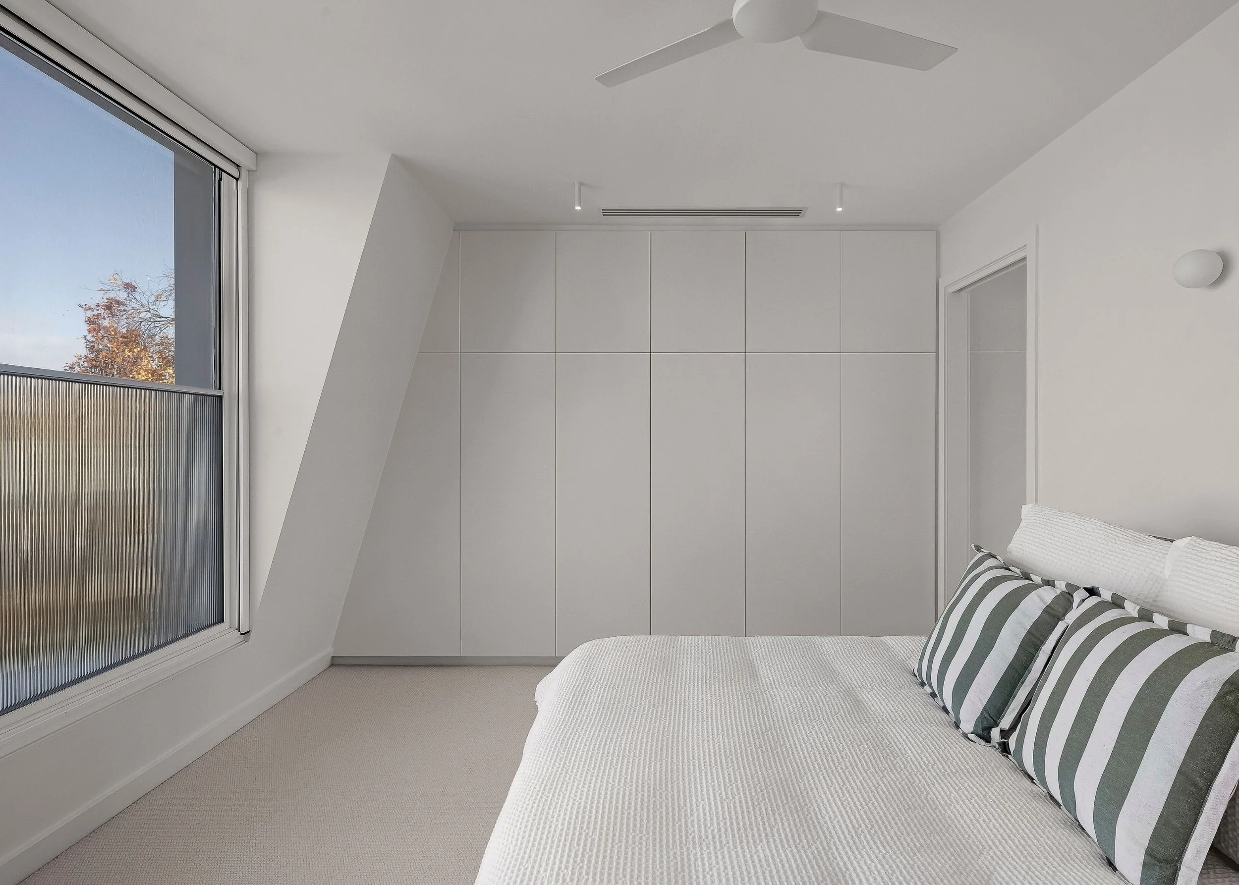 A minimalist bedroom with white walls, a large window showing blue sky and trees, a white ceiling fan, built-in white wardrobe, bed with white bedding and striped pillows, and beige carpet.