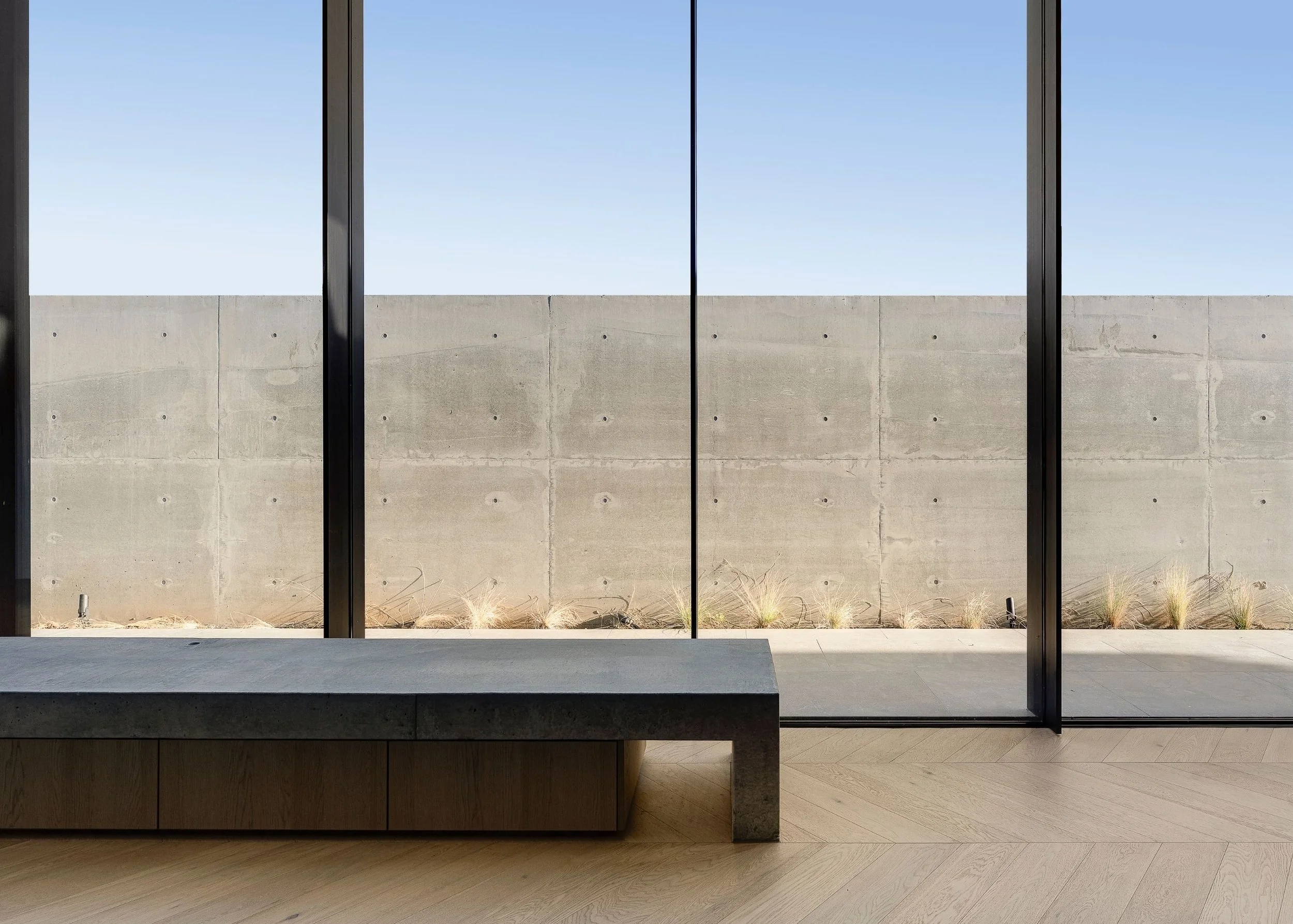 Interior view through large glass window showing a concrete wall, a clear blue sky, and some low desert plants outside. The window has black metal framing and a concrete ledge inside.