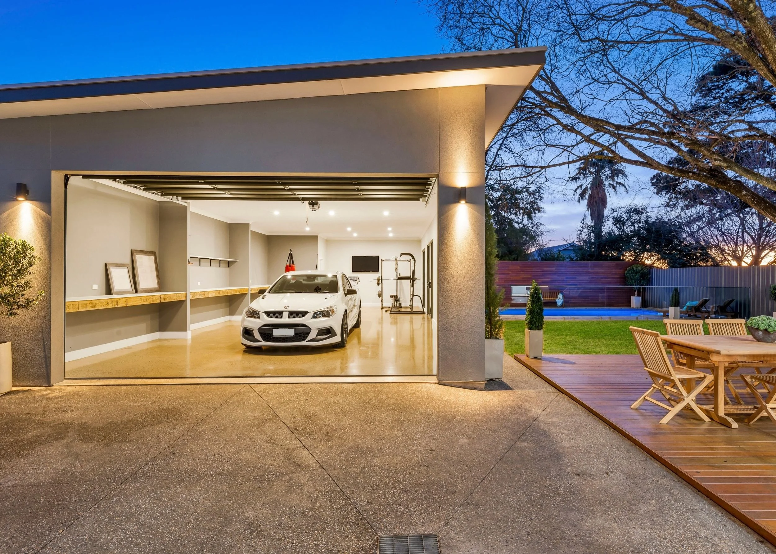 Modern garage with a white car parked inside, open door revealing a neatly organized interior with framed artwork and shelves. Outside, a wooden deck with seating area and outdoor furniture, trees, bushes, and a sunset sky in the background.