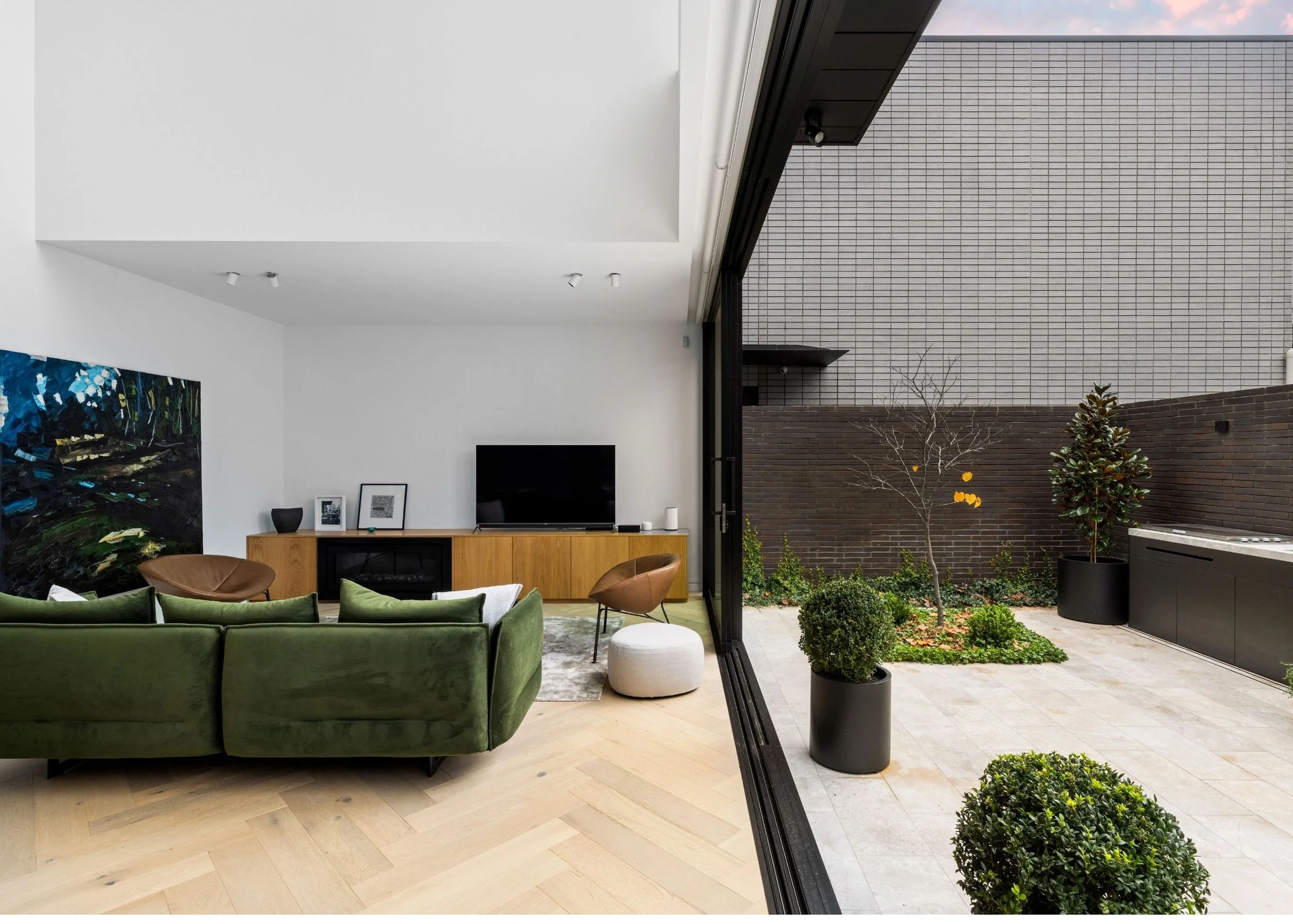 Interior view of a modern living room with a green velvet sofa, wooden cabinet with artwork, and a large open sliding glass door leading to a backyard with potted plants and a brick wall.