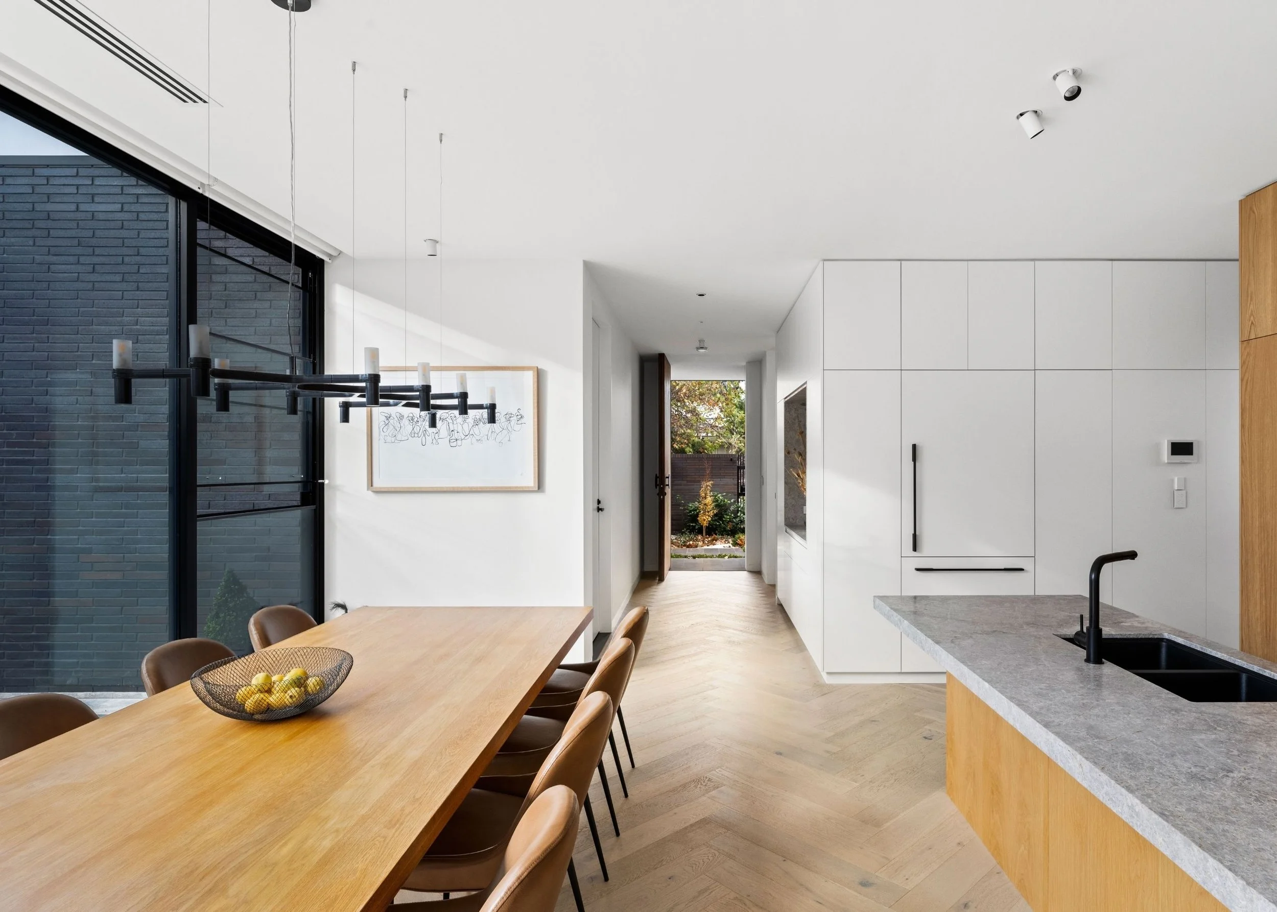 Modern minimalist kitchen and dining area with wooden dining table, brown chairs, white cabinetry, black faucet, and a large window showing greenery outside.