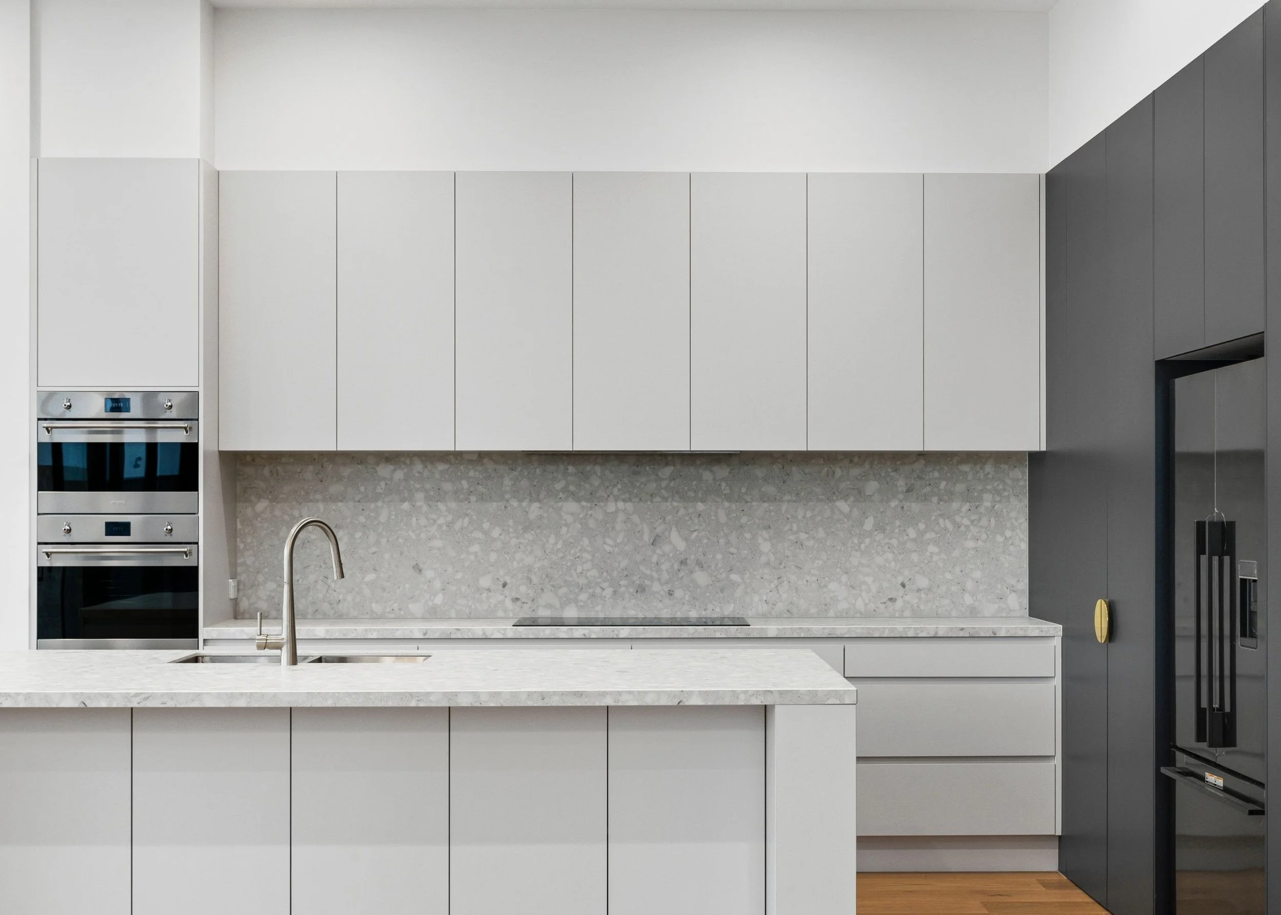 A modern kitchen with white upper cabinets, a gray wall cabinet, a light stone backsplash, a white countertop, a stainless steel oven, and a black refrigerator.