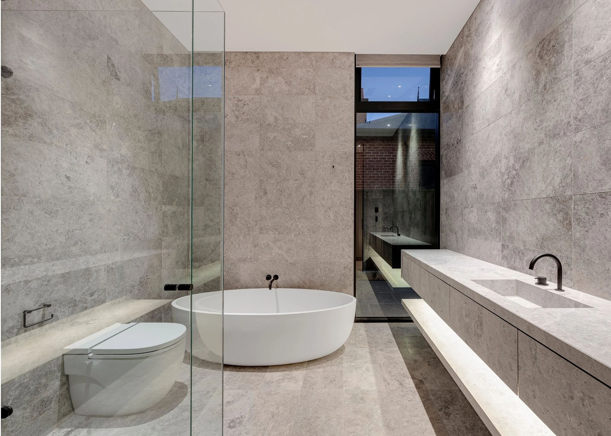 Modern bathroom featuring a freestanding bathtub, a wall-mounted toilet, a vanity with a sink, dark fixtures, and large windows with an outdoor brick wall visible.