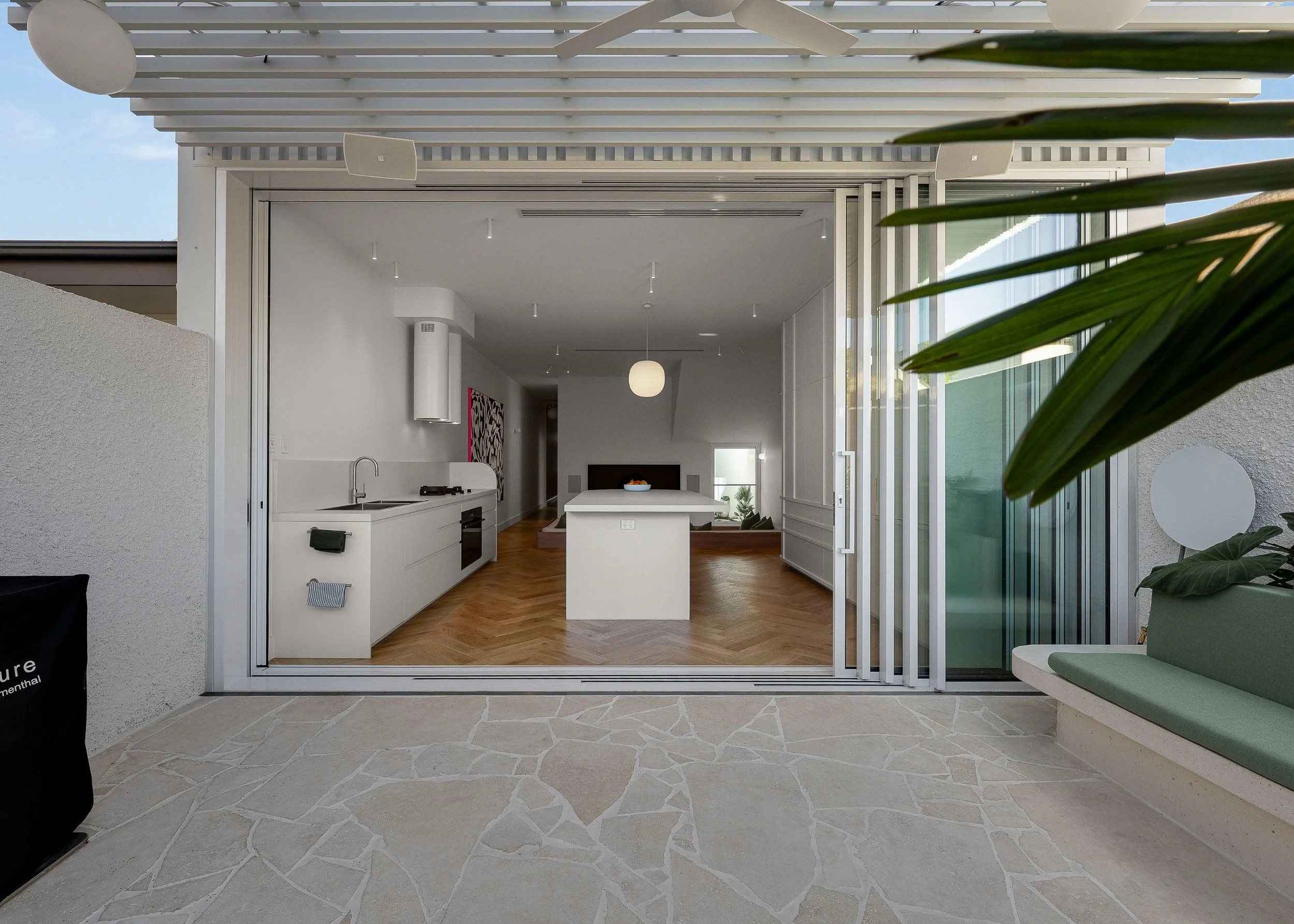Modern interior view of a kitchen and living room area seen through large glass sliding doors, with a green outdoor landscape showing in the background.