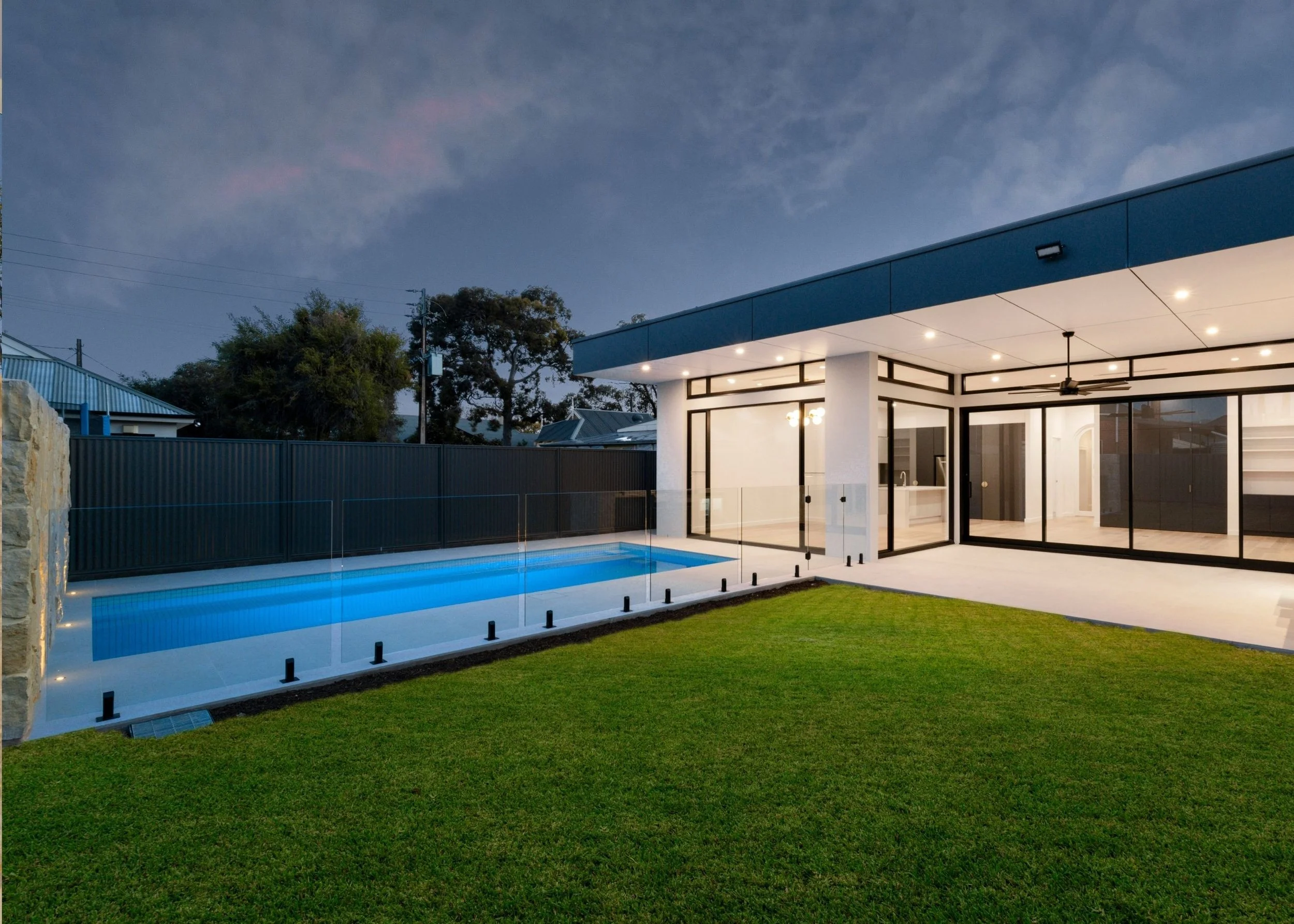Modern house with a glass door, outdoor lighting, and a swimming pool, surrounded by a green lawn at dusk.