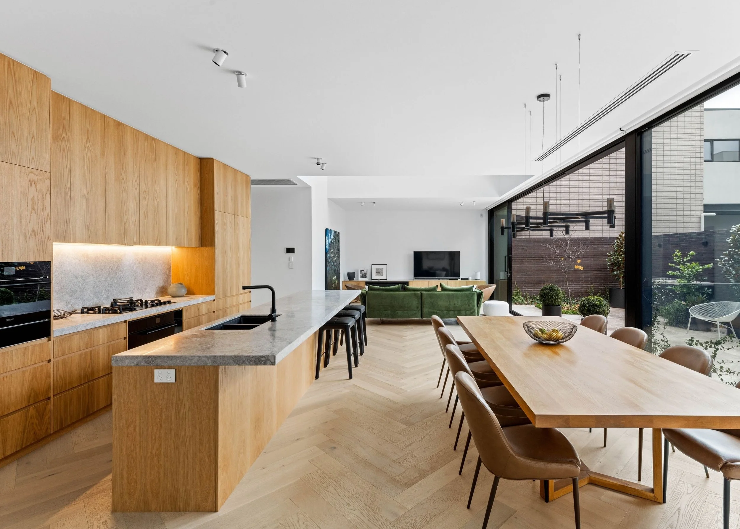 Modern open-plan kitchen and living area with wooden cabinetry, a large kitchen island, a dining table with chairs, a green sofa, and large glass sliding doors that open to an outdoor space with plants and neighboring buildings.