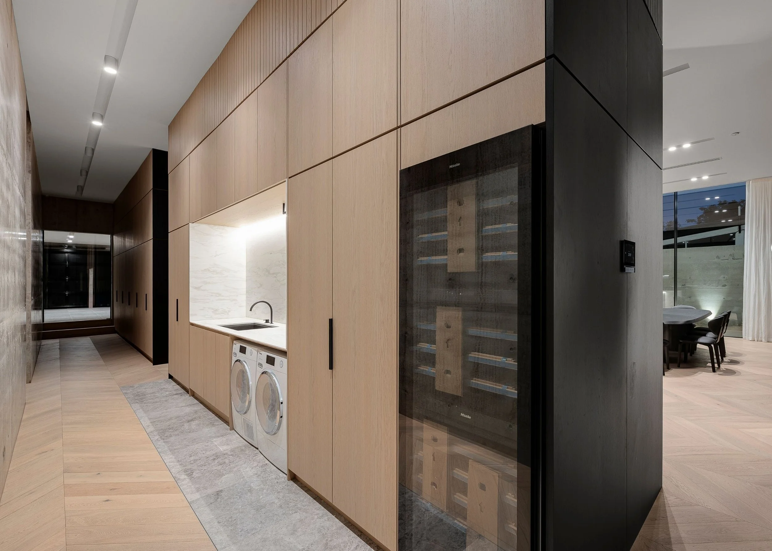 Modern interior kitchen and laundry area with wooden cabinetry, black wine fridge, washing machines, and white marble backsplash.